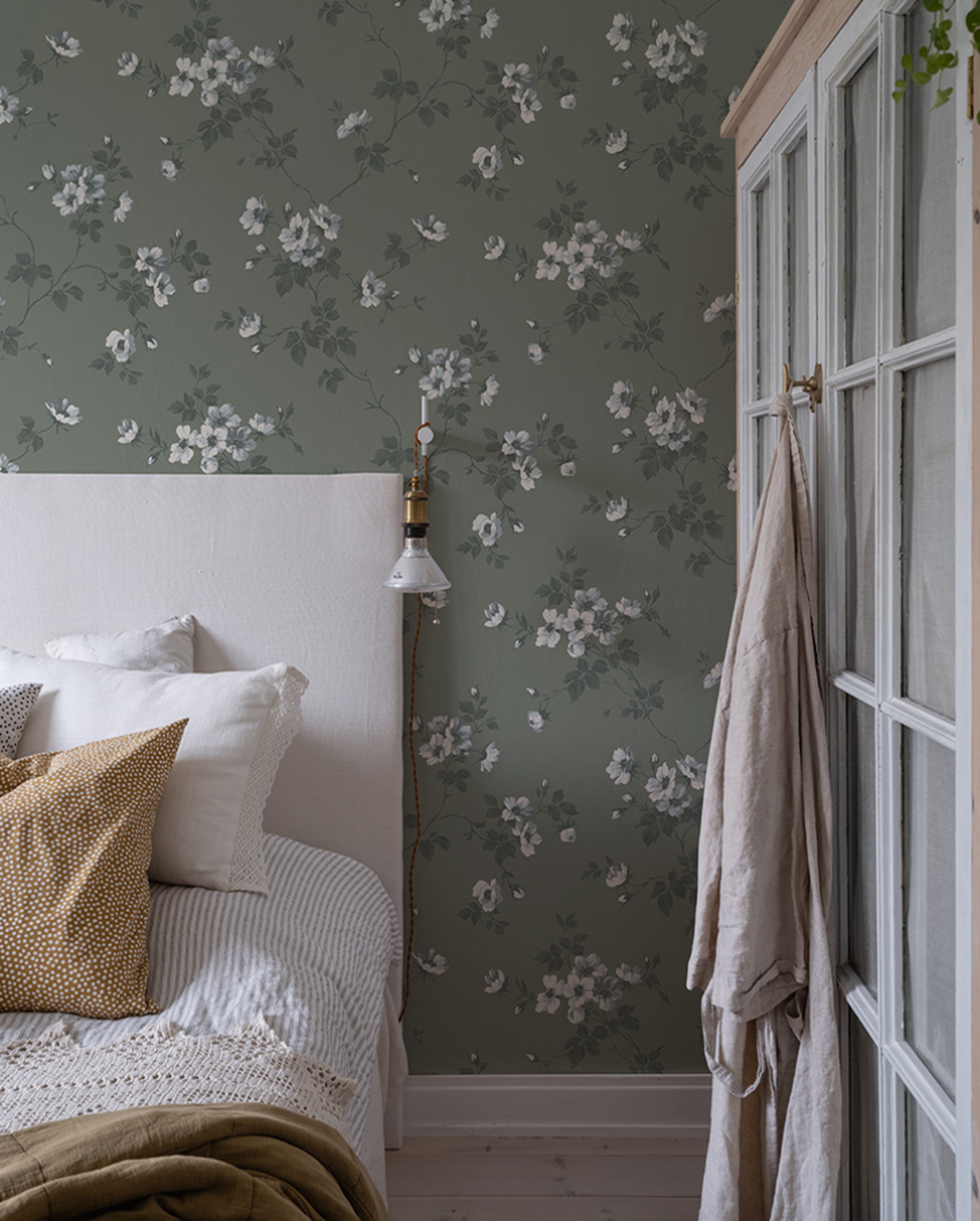 A bedroom with green floral wallpaper, a white headboard, and a wardrobe with a robe hanging on it.