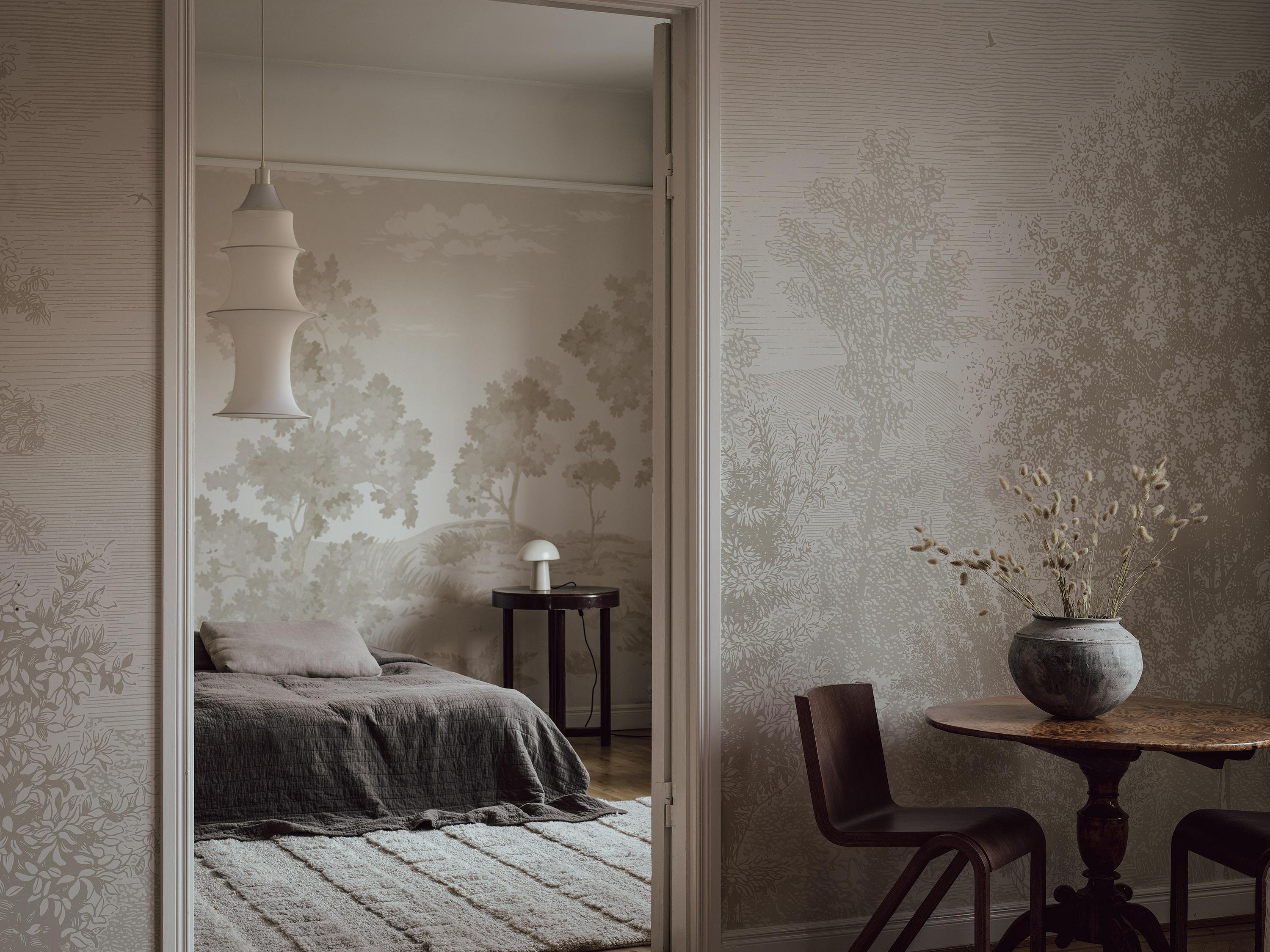Muted, nature-patterned wallpaper covers the walls of a bedroom with a bed and lamp, seen through a doorway from an adjacent room with a table and dried flowers.