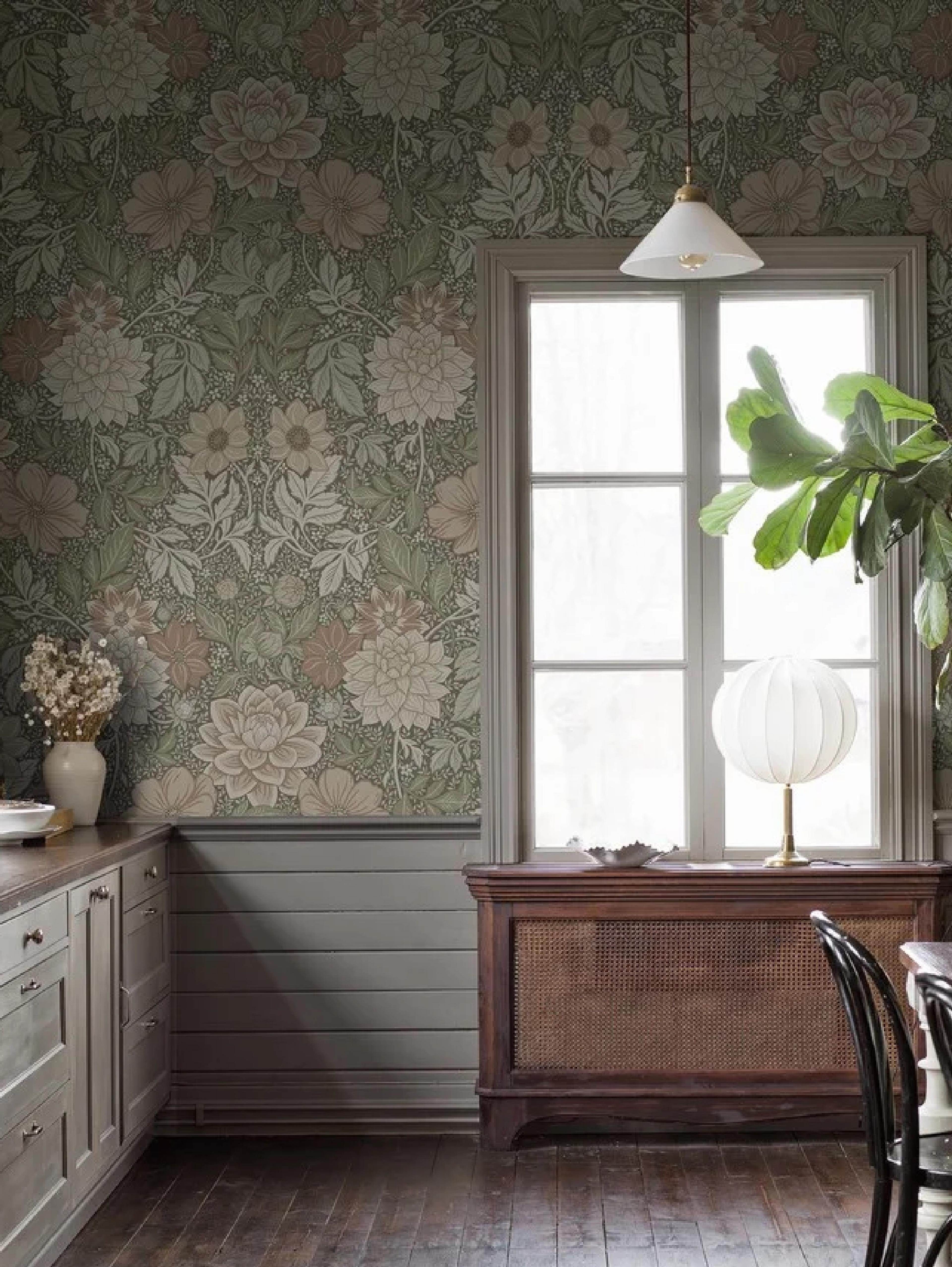 A room with green floral wallpaper, light cabinets, a large window over a dark wood cane cabinet, and dark wood floors.