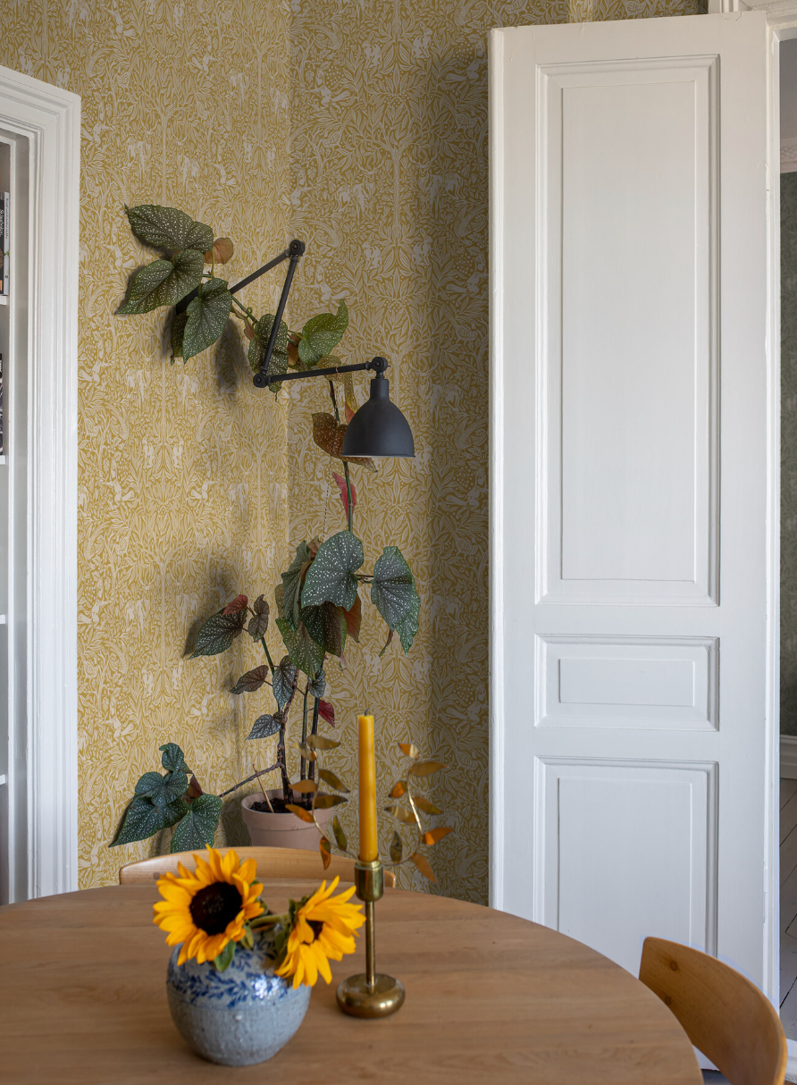 Yellow patterned wallpaper covers a room with a tall plant under a black wall lamp. A wooden table in the foreground holds a vase of sunflowers and a yellow candle.