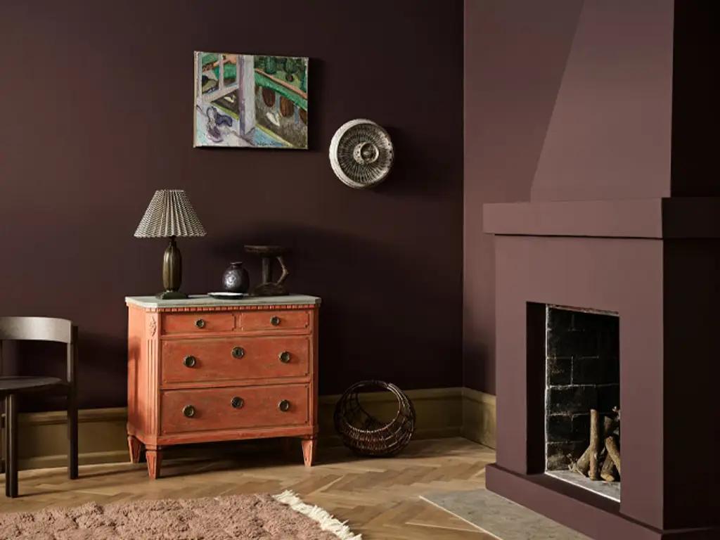 A room with deep plum walls, a matching modern fireplace, a coral antique dresser with a lamp, and herringbone wood floors.