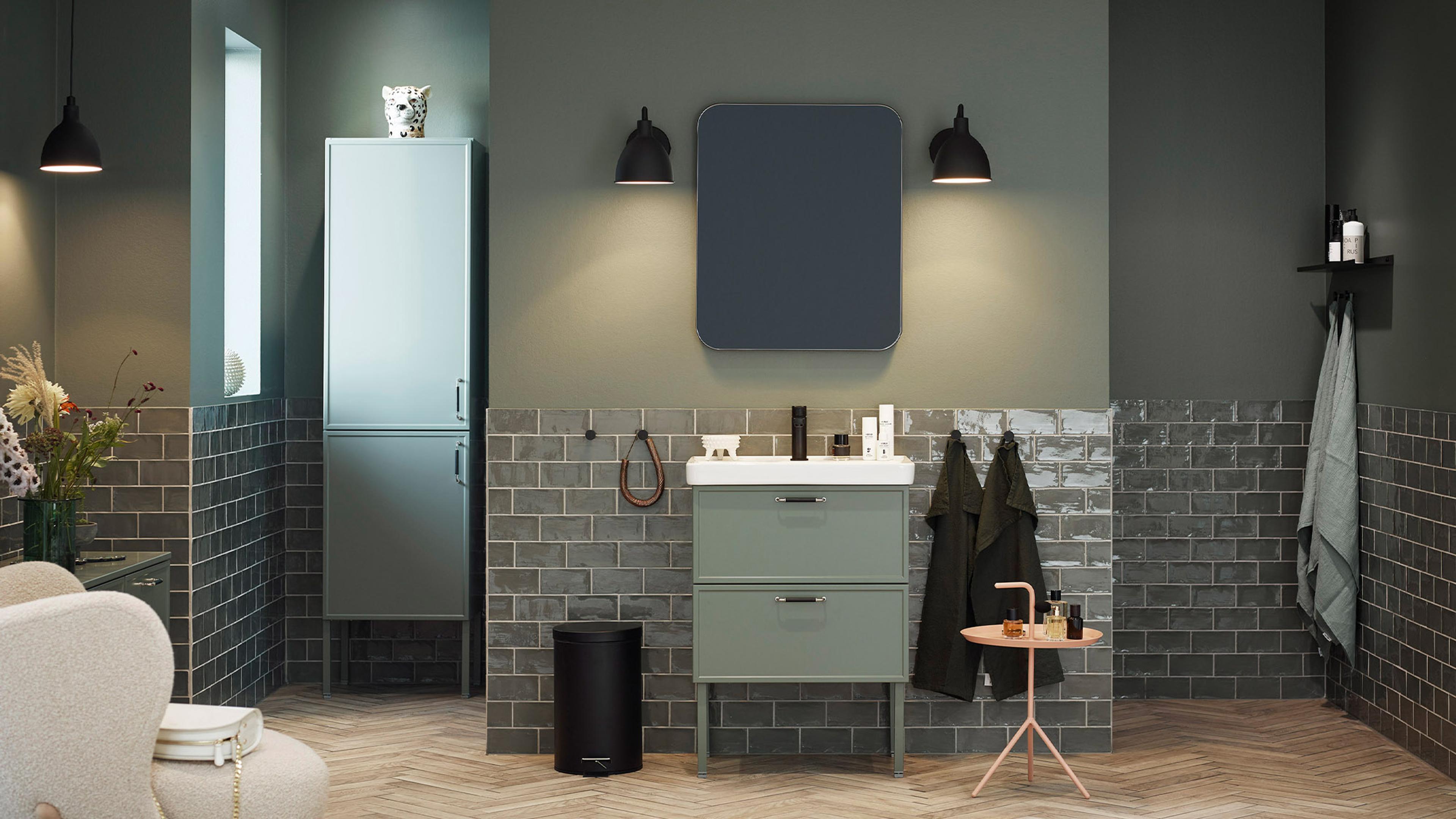 Stylish modern bathroom with olive green walls, dark gray subway tiles, a light green vanity with sink, tall cabinet, dark mirror, and herringbone wood floor.