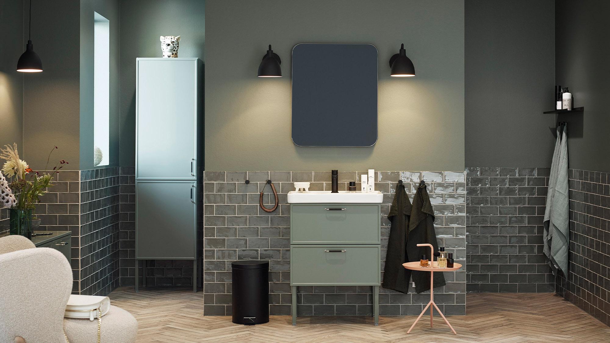 Stylish modern bathroom with olive green walls, dark gray subway tiles, a light green vanity with sink, tall cabinet, dark mirror, and herringbone wood floor.