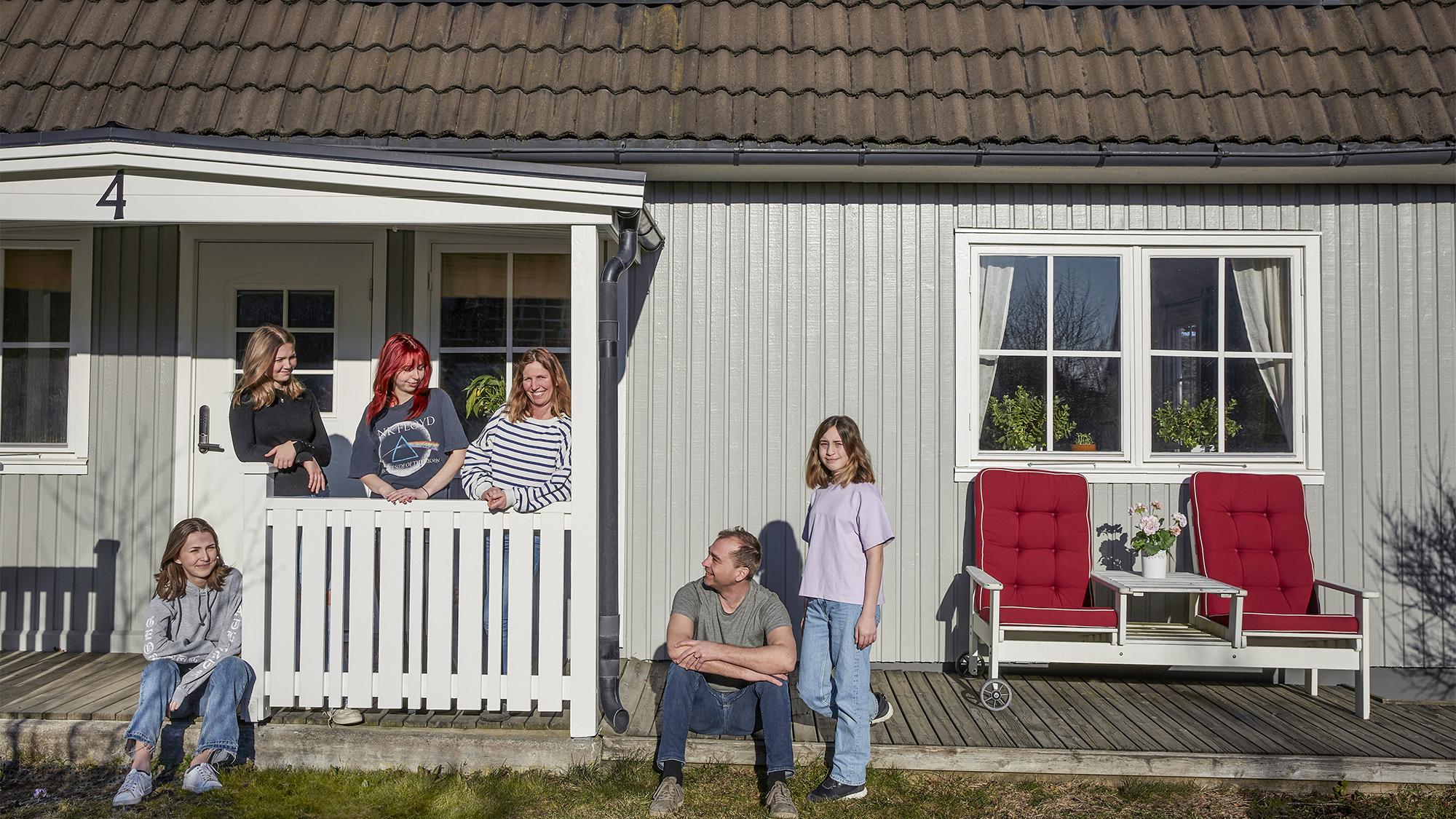 A family of six poses outside a light grey house with a porch and wooden deck.
