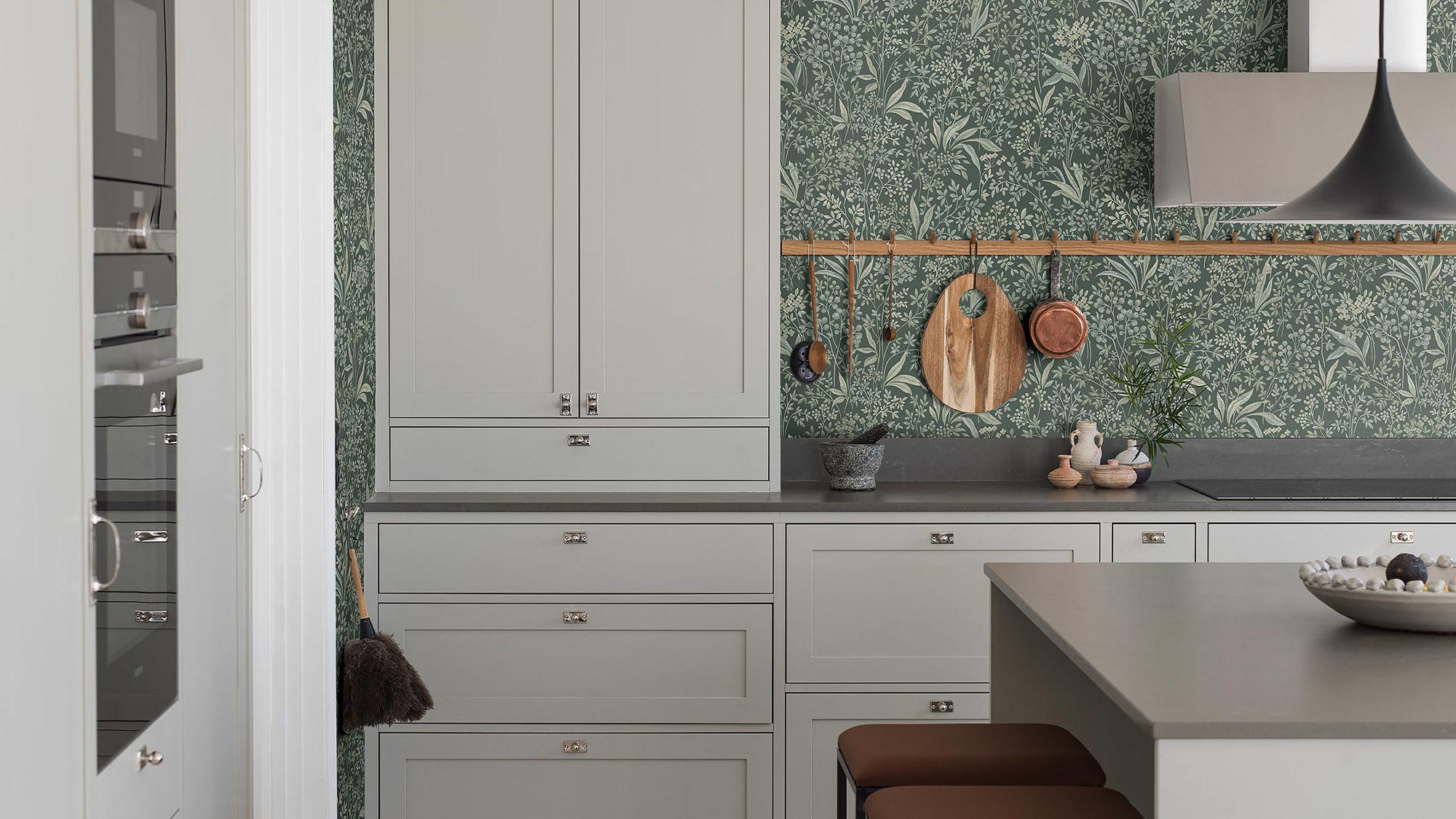 A modern kitchen with light gray cabinets, dark countertops, and green botanical wallpaper, featuring a wooden rail with hanging utensils and a kitchen island.