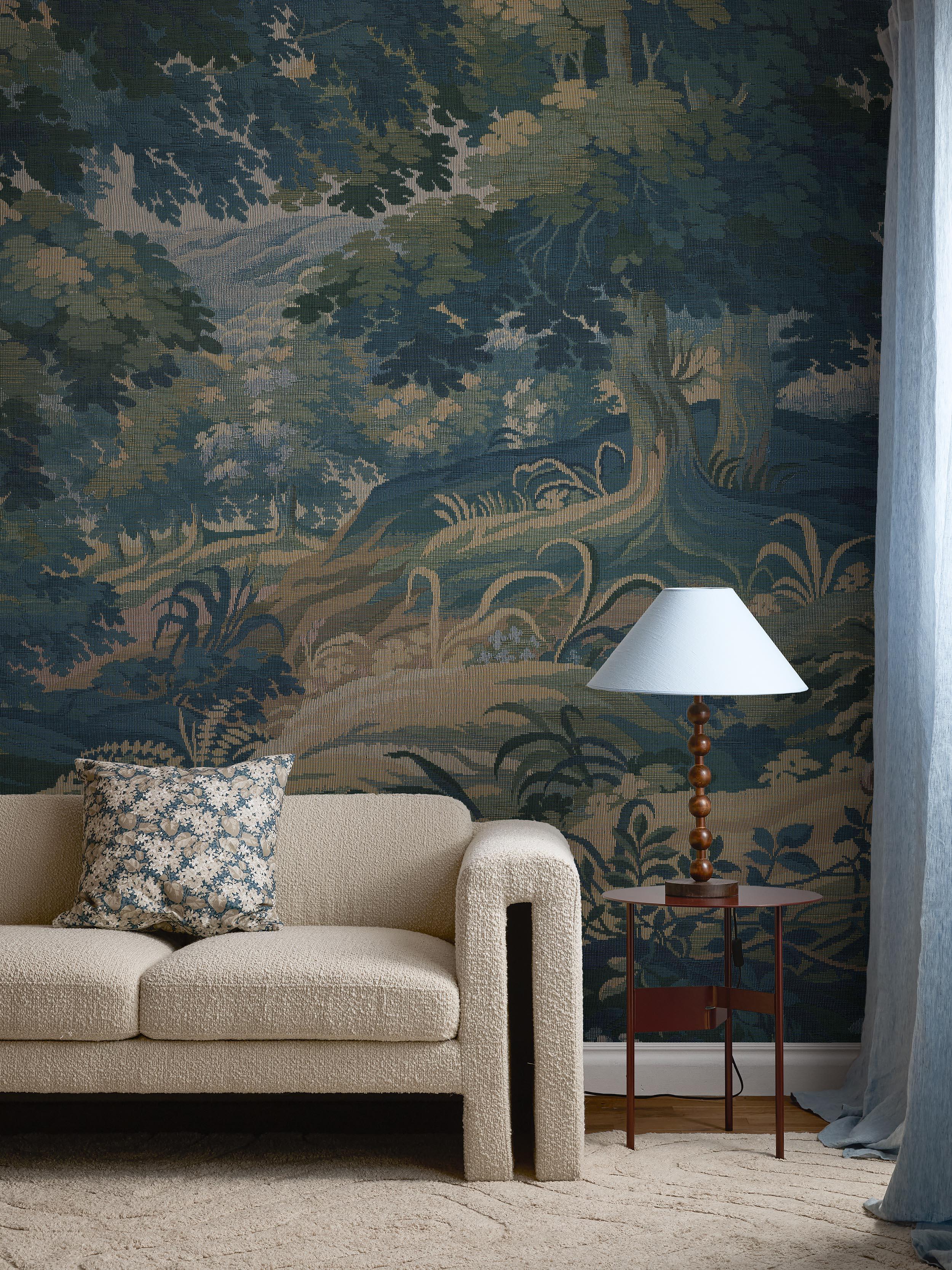 A room with a blue-green forest tapestry wallpaper, a cream textured sofa with a patterned pillow, and a wooden lamp on a side table.