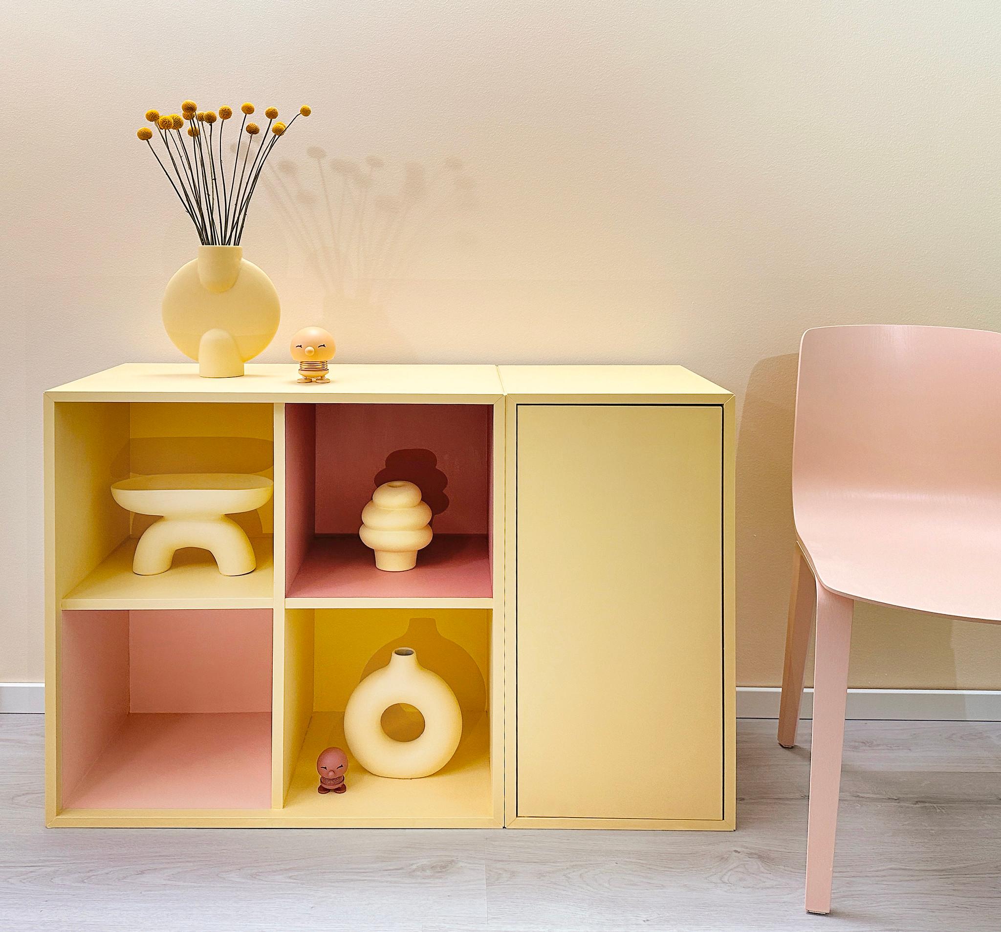 A light yellow modular cabinet with pink and yellow cubbies displays various pale yellow sculptural items, next to a matching pink chair.