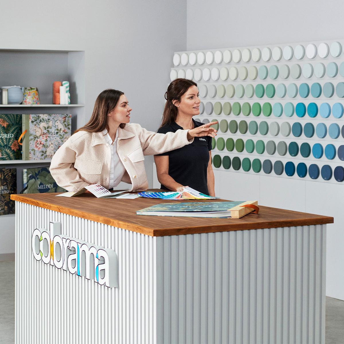 Two women at a Colorama store, one pointing at a large wall display of paint color samples.