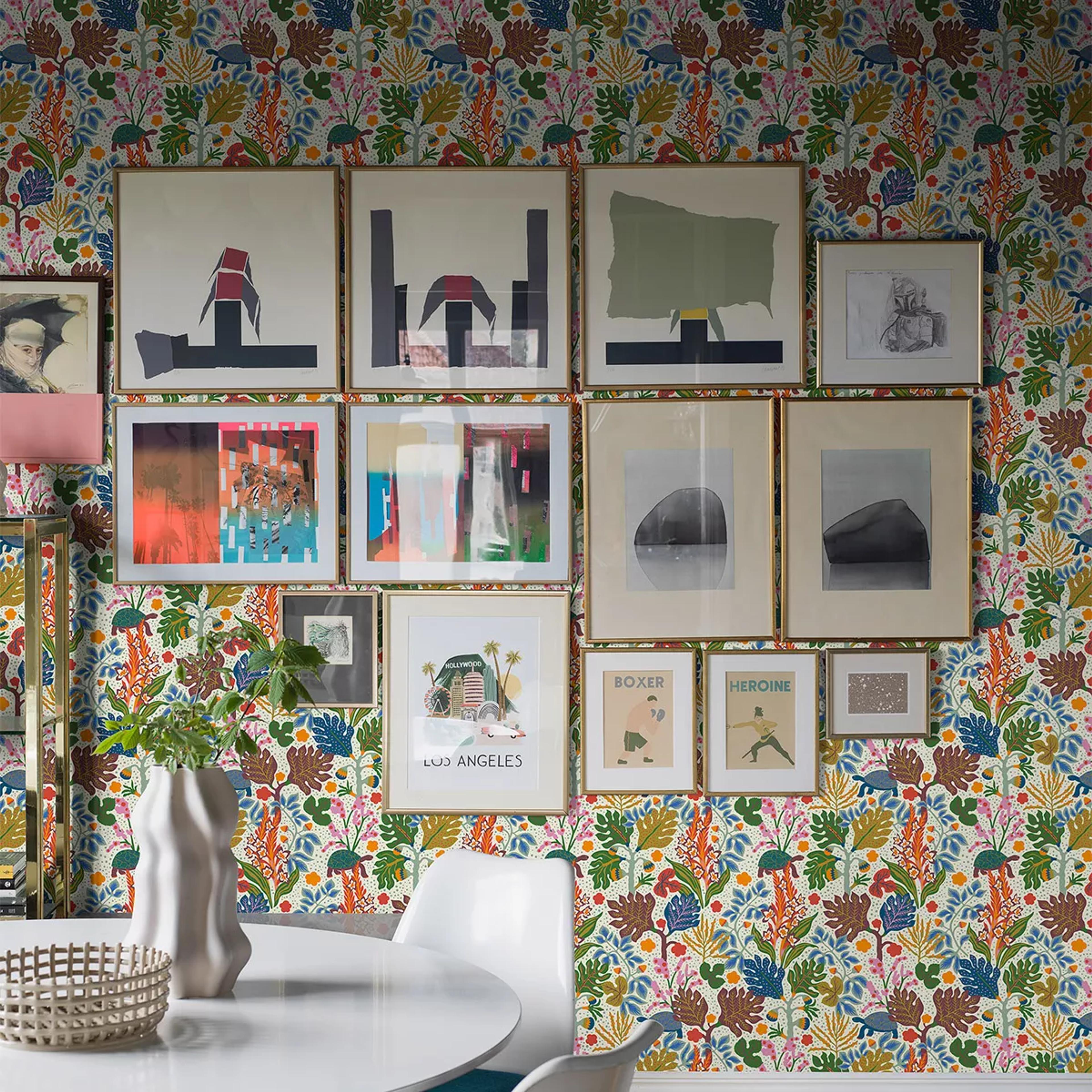 A room with vibrant botanical wallpaper, a large gallery wall of various framed artworks, and a white dining table with chairs in the foreground.