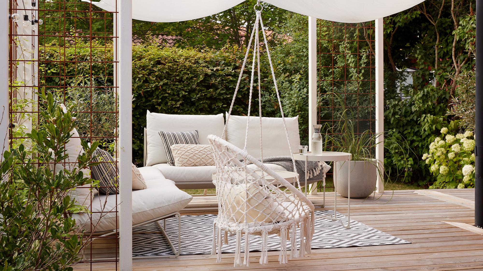 An outdoor patio with a wooden deck, white furniture including a sectional and hanging macrame chair under a white canopy, surrounded by garden foliage.