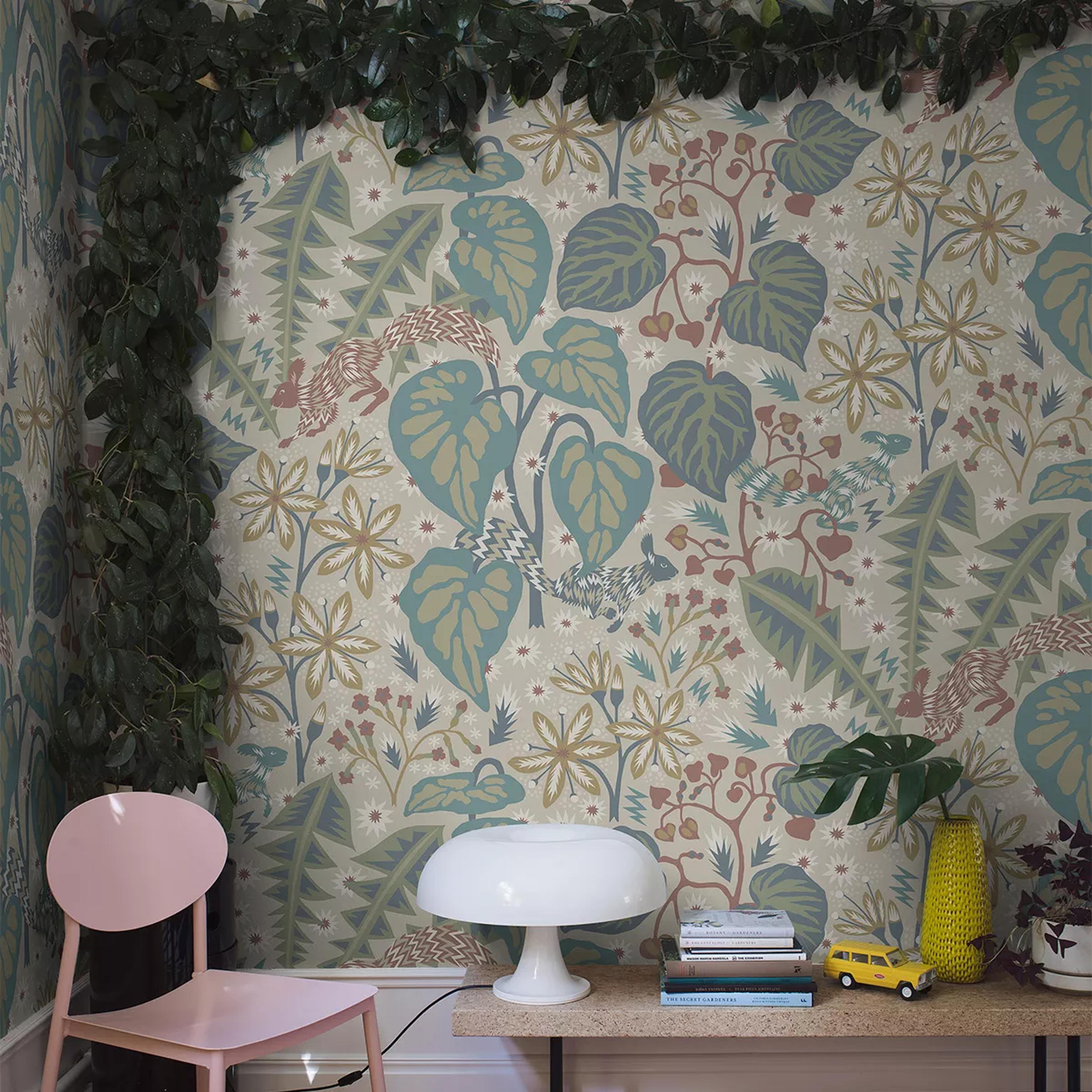 A room with beige nature-patterned wallpaper featuring blue-green leaves, rust flora, and squirrels, framed by a green vine, and a pink chair next to a table with a white mushroom lamp.