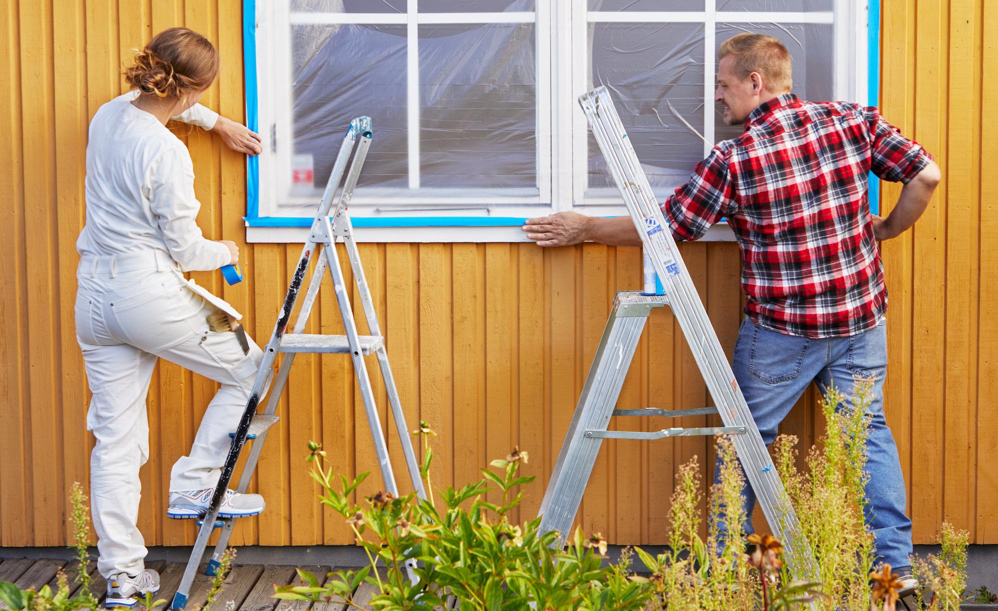 A man and woman tape around a house window, preparing it for painting.