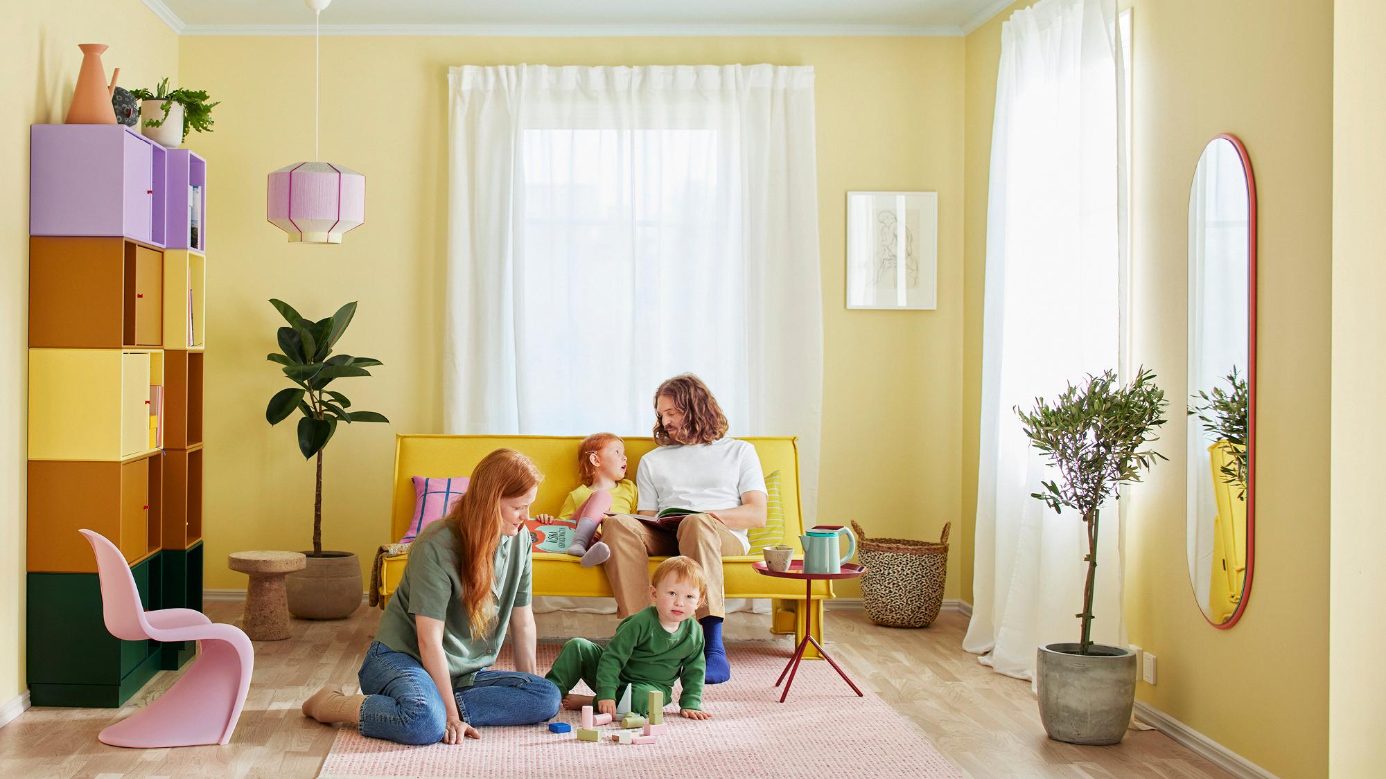 A family of four in a yellow-walled living room: a mother plays with a toddler on a rug, and a father reads to a girl on a yellow sofa.