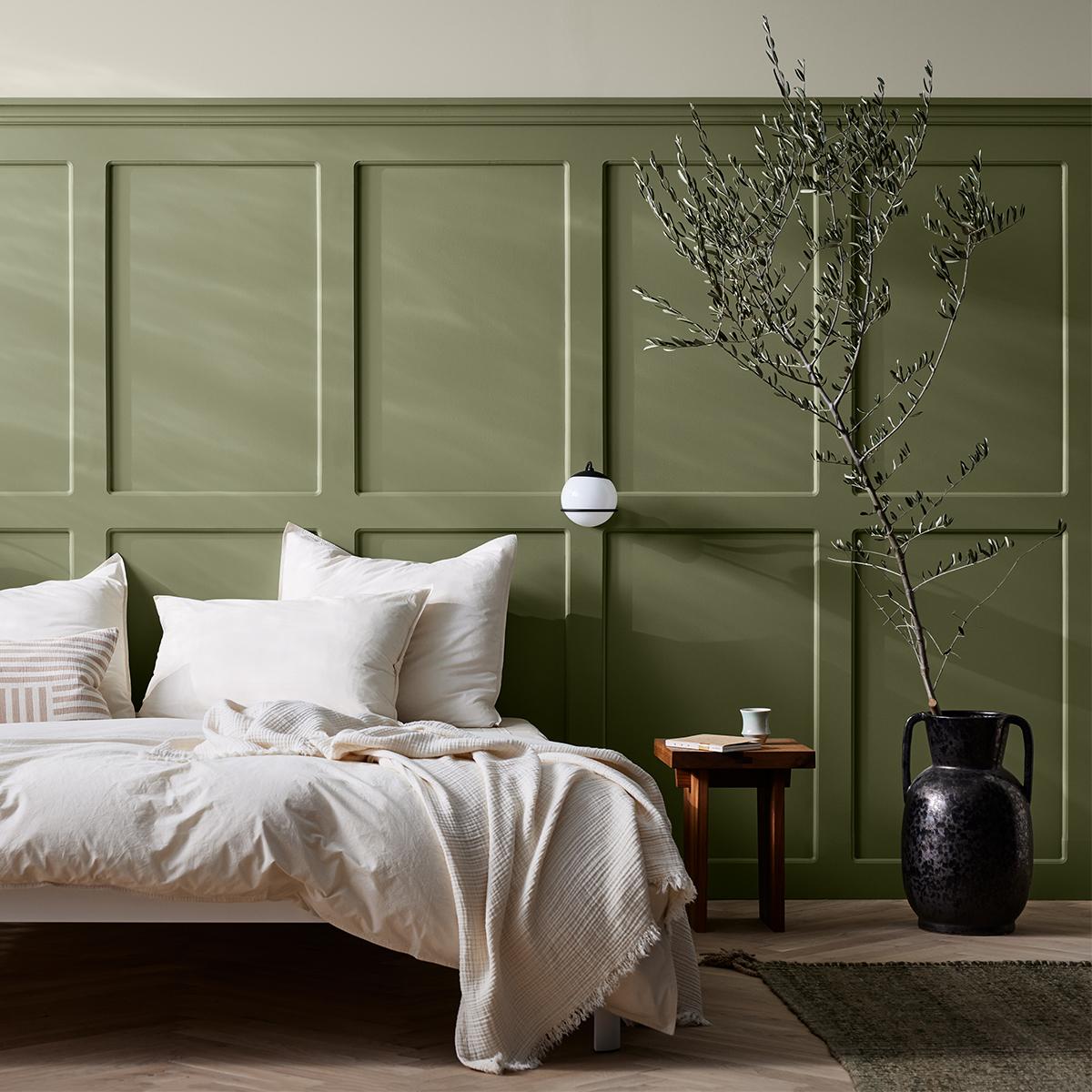 A bedroom featuring a sage green paneled accent wall, a bed with white bedding, and a tall olive branch in a dark vase.