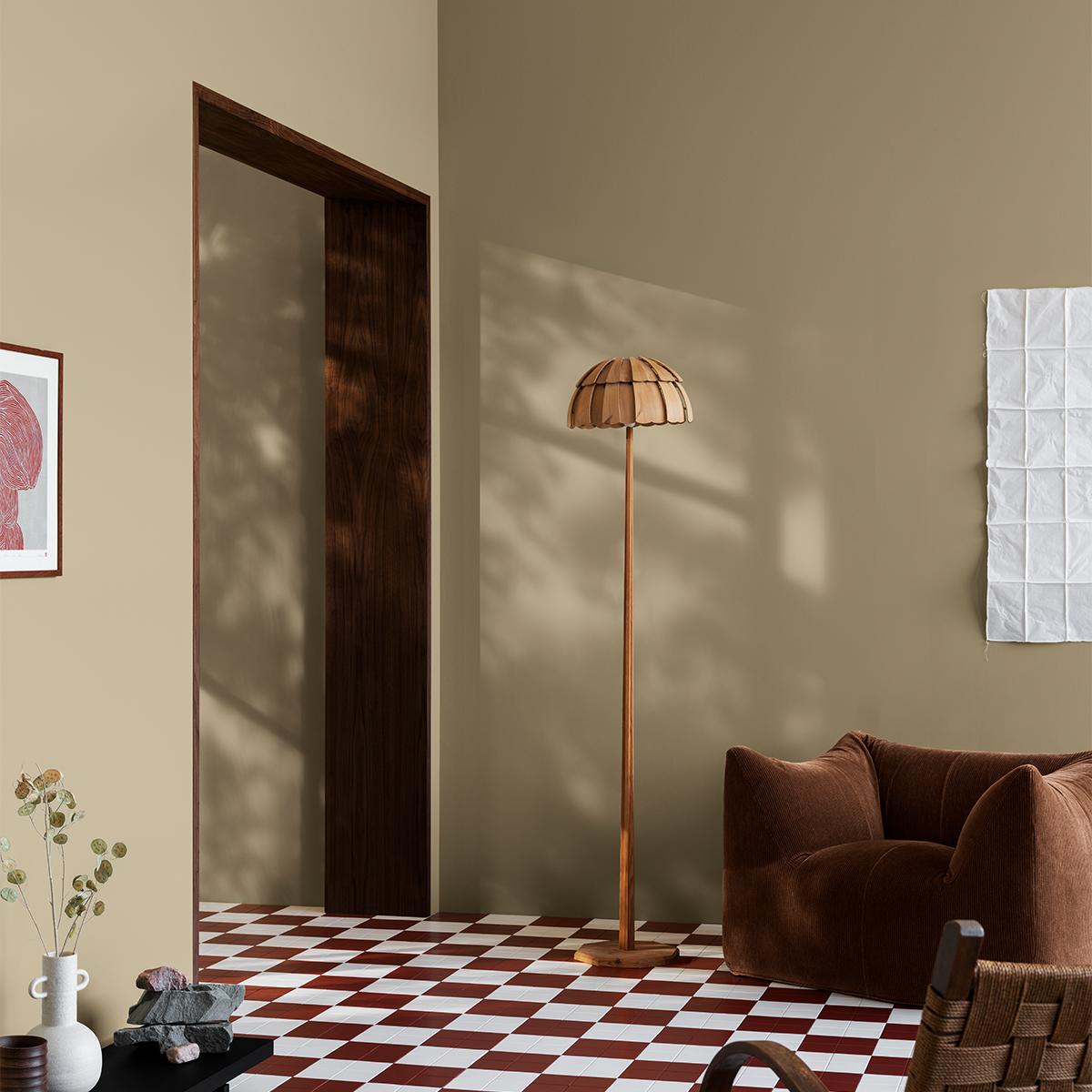 An inviting, earthy-toned room with a red and white checkered floor, a wooden floor lamp, a plush brown armchair, and sunlight casting tree shadows on the walls.