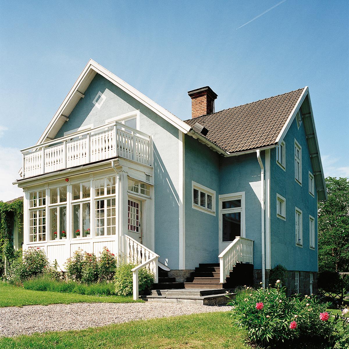 Light blue house with a white balcony, sunroom, and brown roof in a sunny garden.