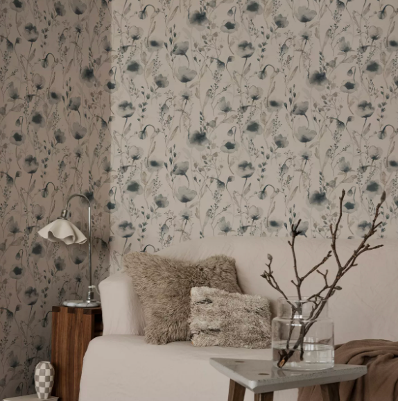 A cozy room featuring light wallpaper with a pattern of blue-grey watercolor poppies and foliage, a bed with fluffy pillows, a lamp, and decorative branches.