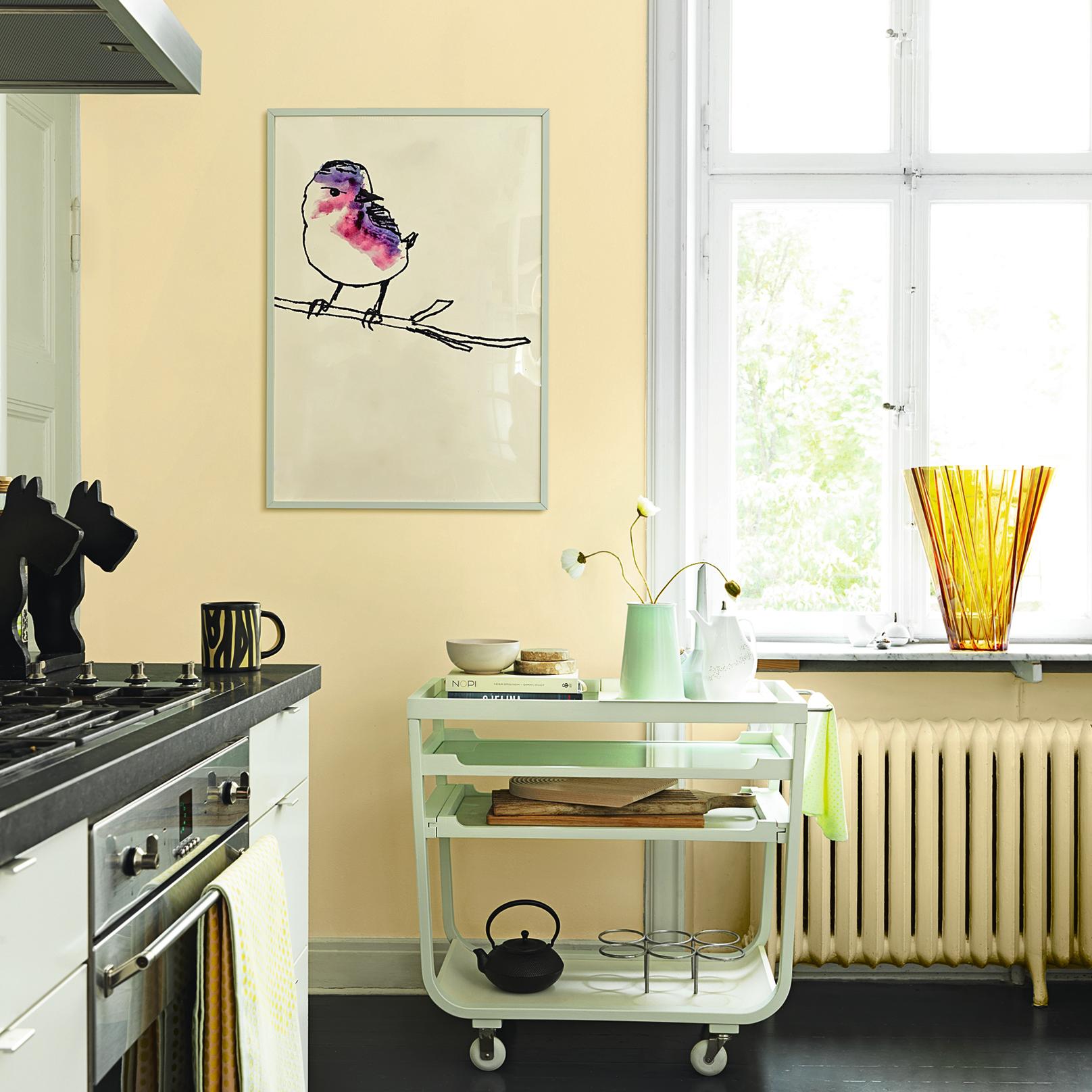 A kitchen with light yellow walls, featuring a framed bird drawing, a white rolling cart, and a large window.
