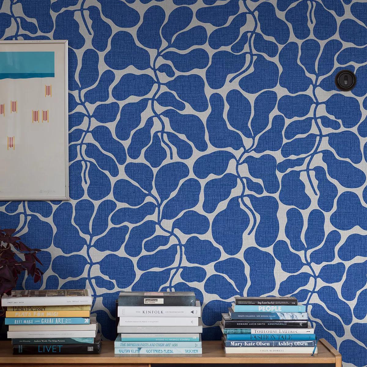 Blue and white abstract leaf wallpaper, with a framed beach art print and stacks of books on a wooden surface.