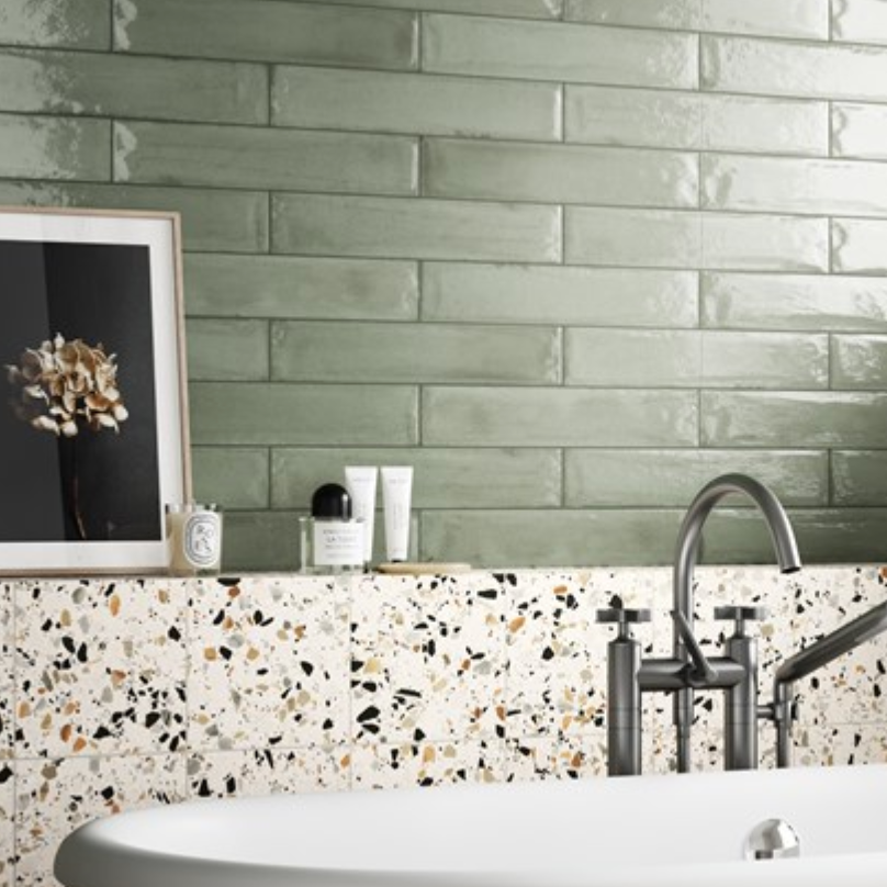 Modern bathroom with a white tub, terrazzo tiles, glossy sage green subway tiles, and a dark metal faucet.