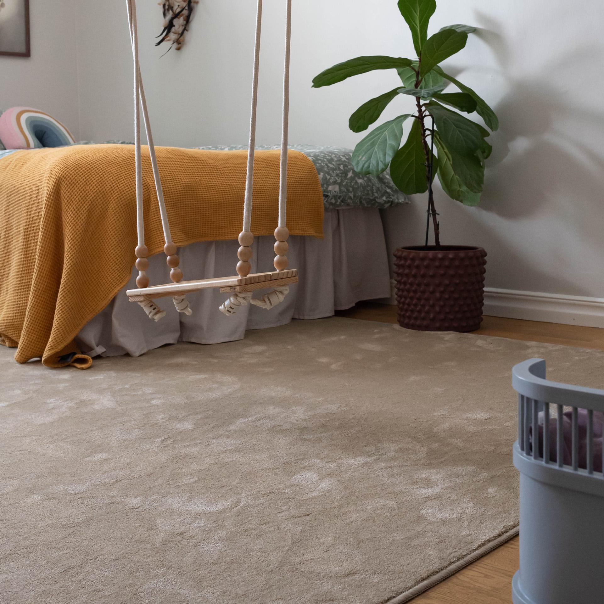 A child's room with a wooden swing, a bed with a yellow throw, a fiddle leaf fig plant, and a large beige rug.