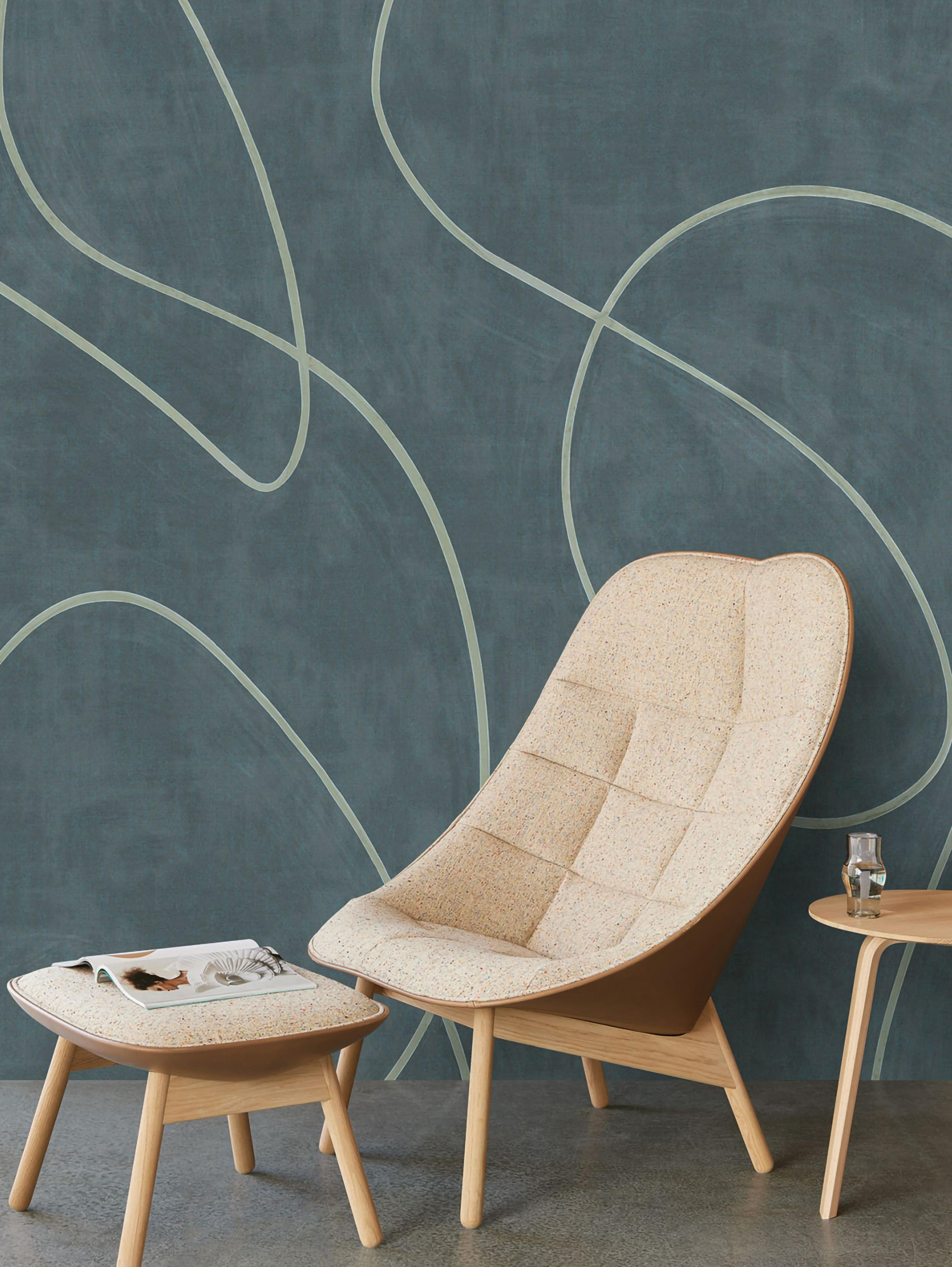 A modern living space with a quilted beige armchair and ottoman, a wooden side table, and a teal wall with abstract lines.