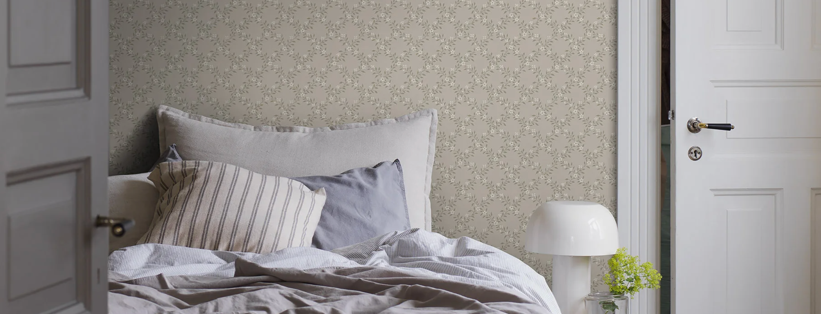 A bedroom with a bed, white lamp, and walls covered in light beige wallpaper with a delicate floral pattern.