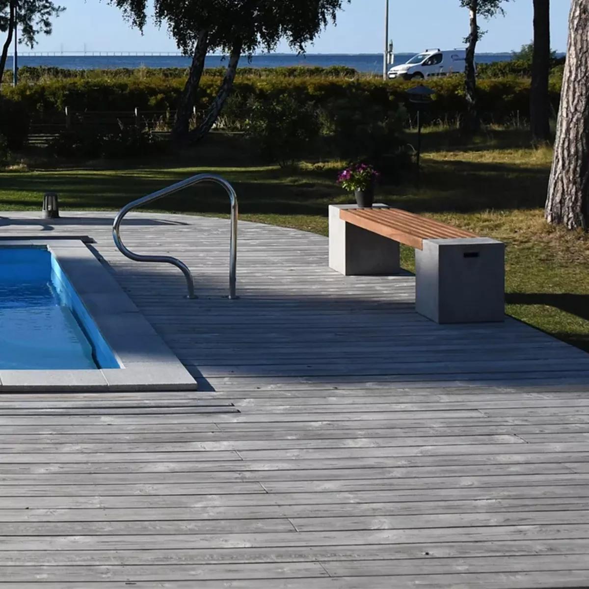 Modern swimming pool with a handrail alongside a wooden deck, a bench with concrete supports and a wooden seat topped with purple flowers, and trees and water in the background.