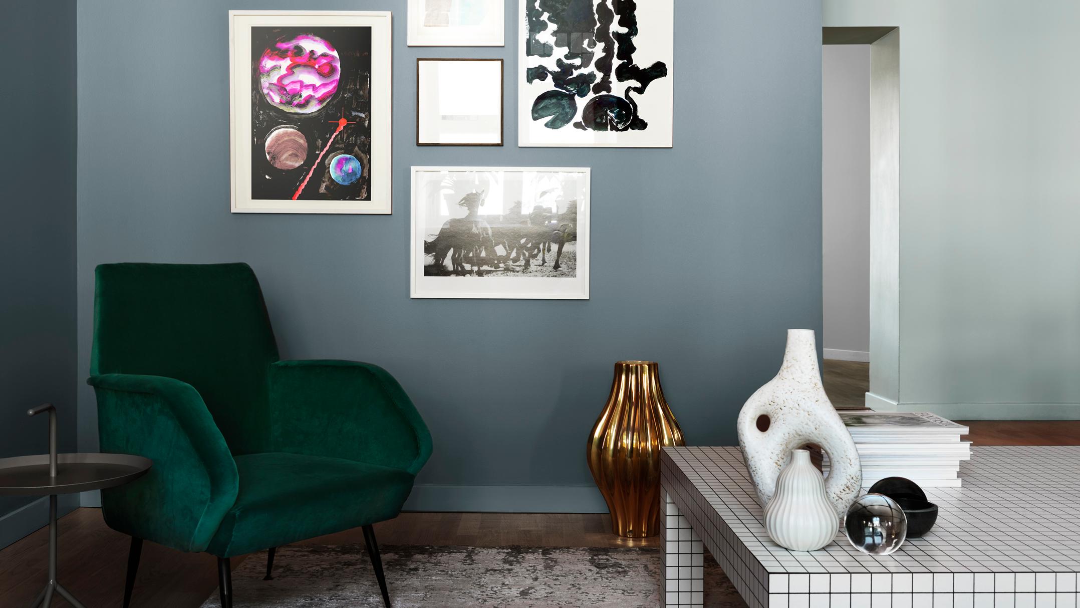 A stylish room featuring a dark green velvet armchair, a gallery wall on a blue-gray wall, and a modern white grid coffee table with decorative vases.