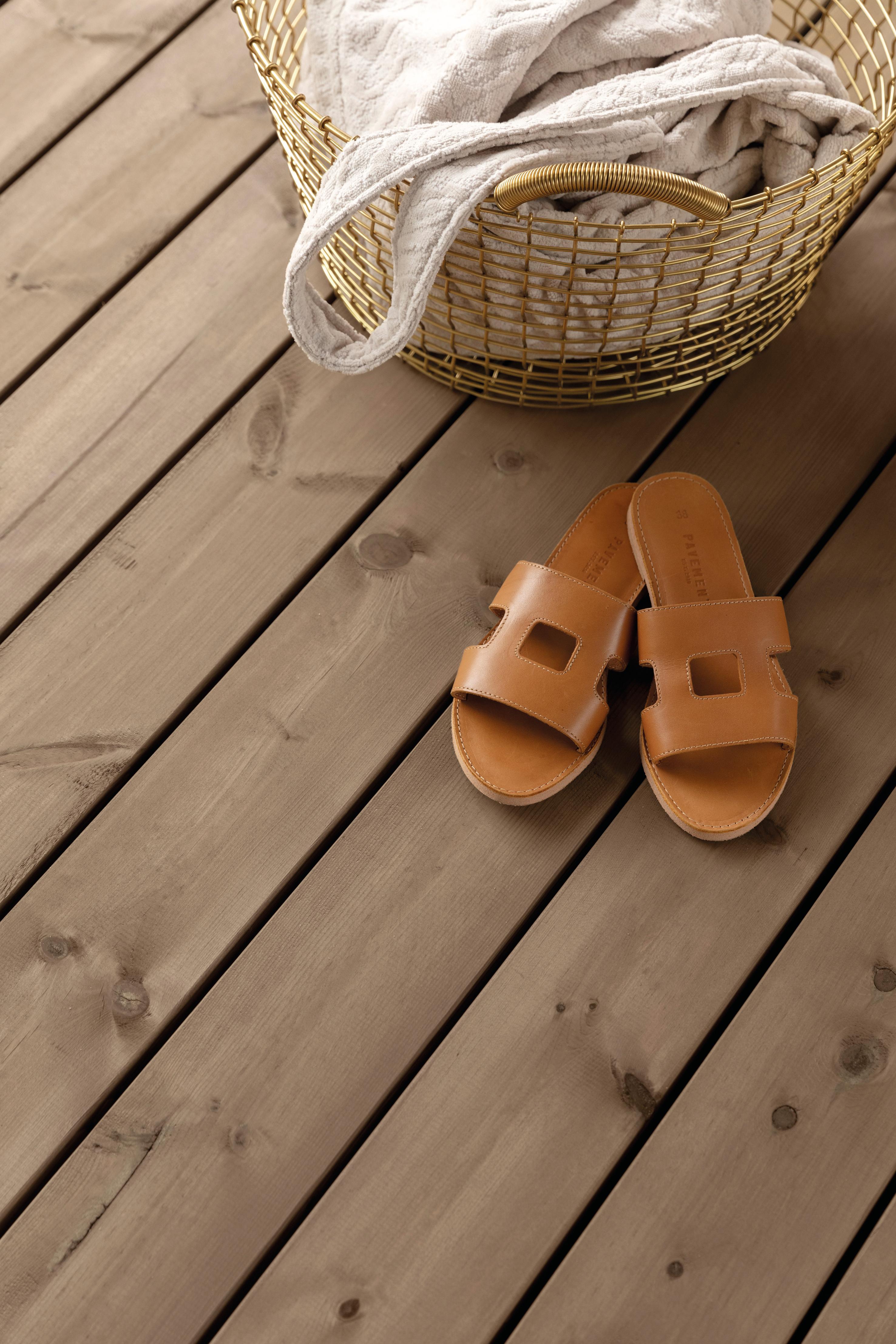 A golden wire basket with a light blanket and brown leather slide sandals on a wooden deck.