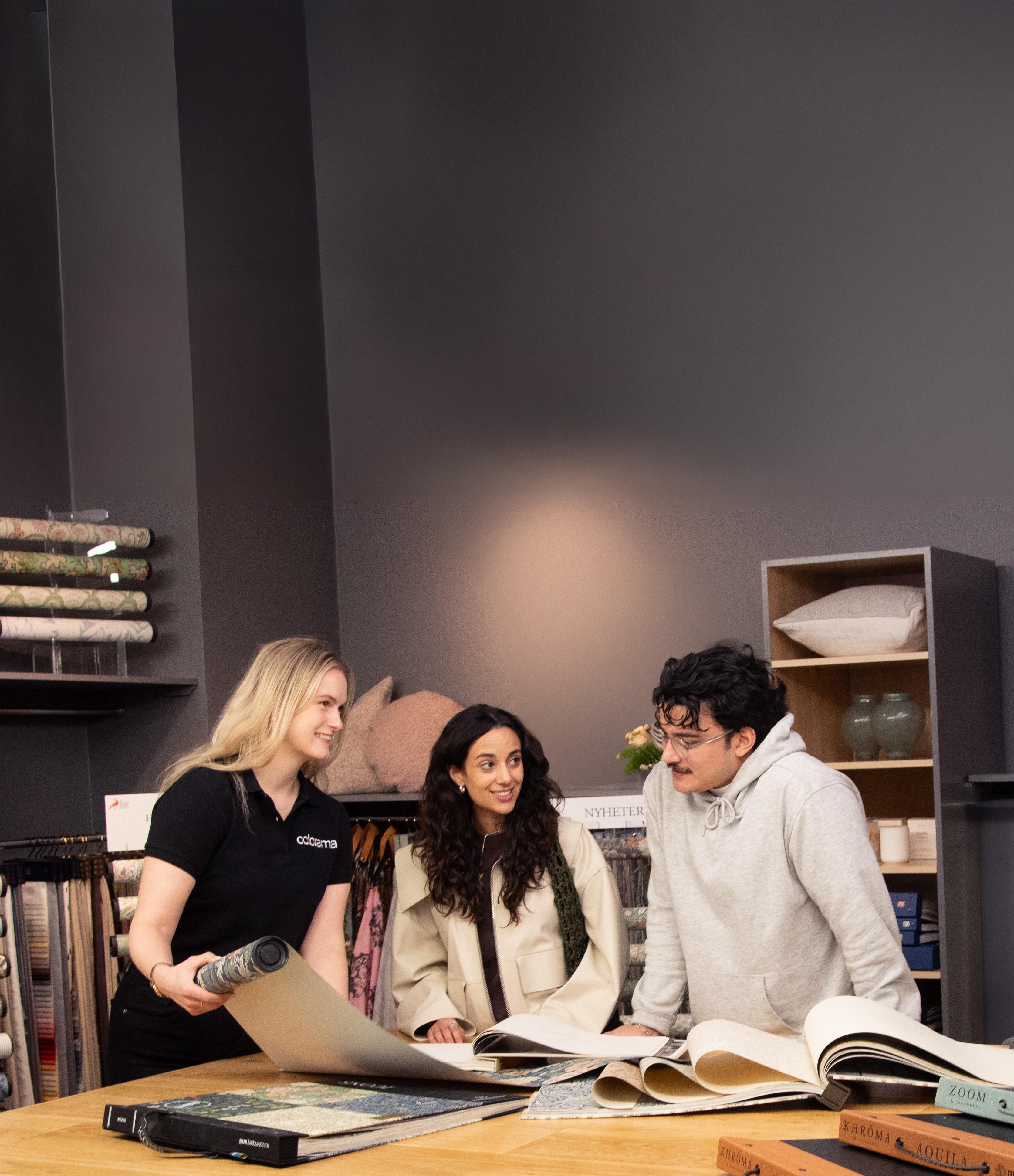 An employee assists two customers browsing wallpaper and fabric samples in a design store.