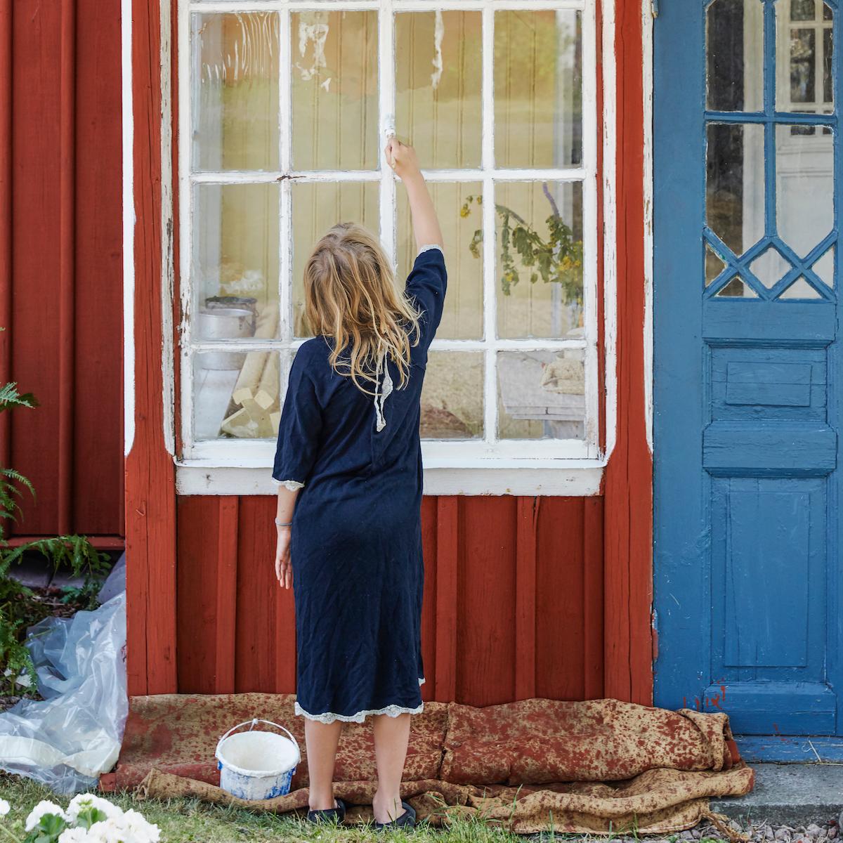 A young girl, seen from behind, paints the white-framed window of a red wooden house.