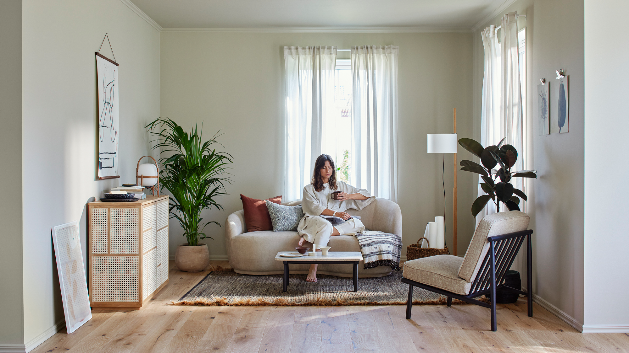 A woman sits on a light sofa in a bright, modern living room with plants.