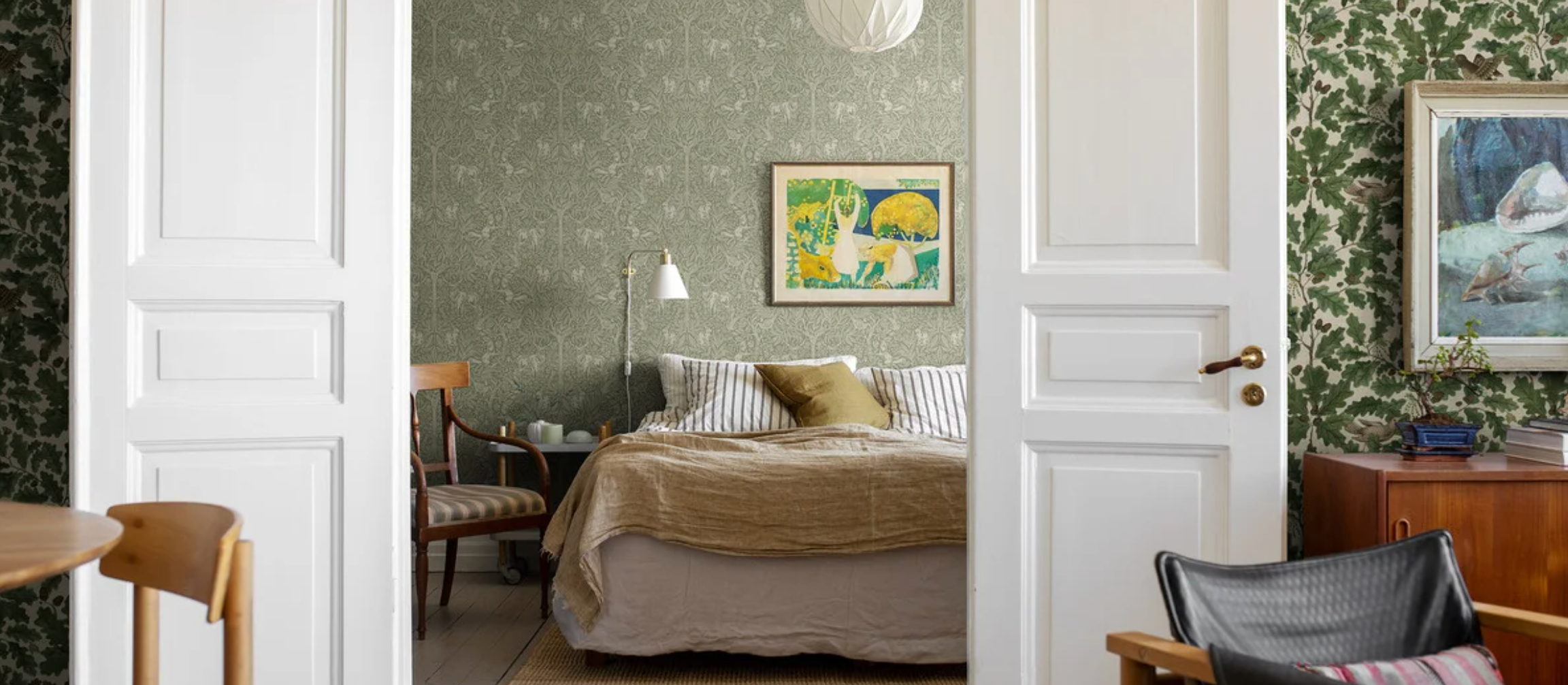 A bedroom with light green patterned wallpaper and a bed, viewed through open white doors from a room with dark green leaf wallpaper.