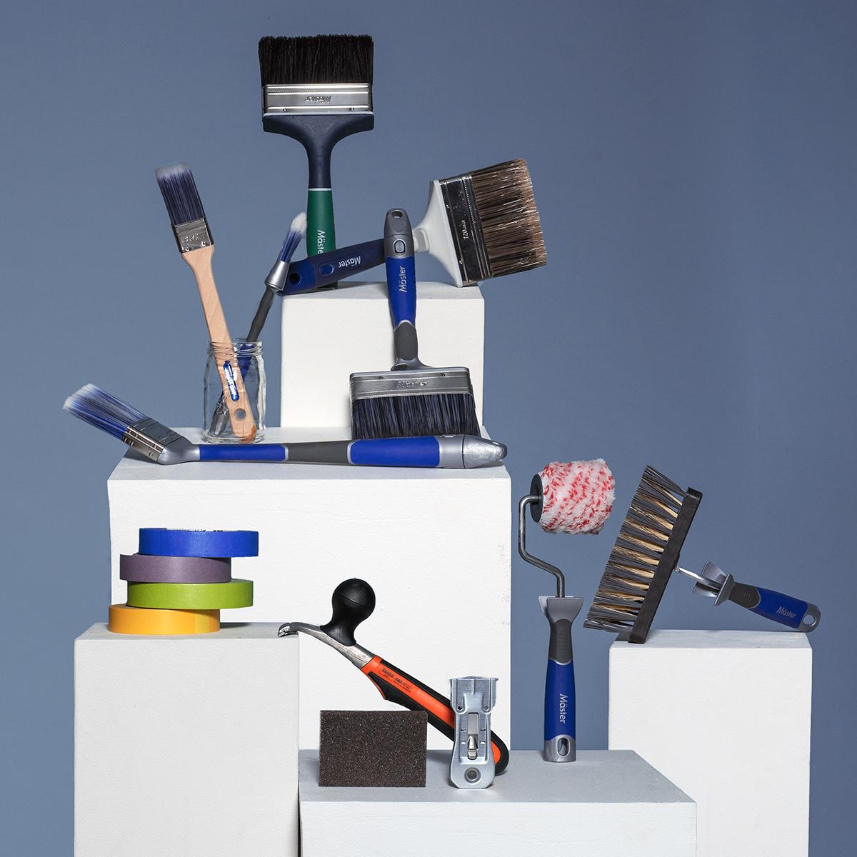 Various painting tools including brushes, a roller, wire brush, multicolored painter's tape, a hammer, sanding sponge, and a scraper, displayed on white blocks.