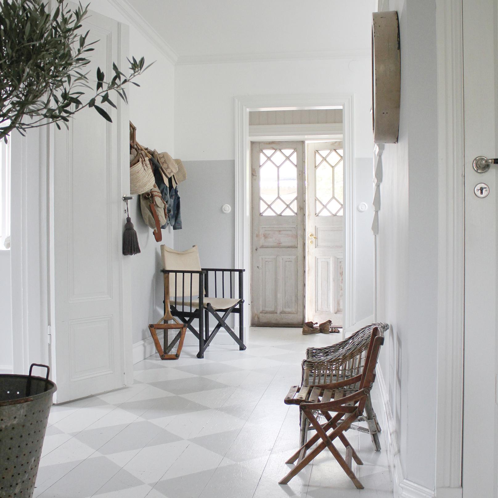 A bright, airy hallway with white and light gray walls, a checkered floor, various chairs, and decorative double doors at the end.