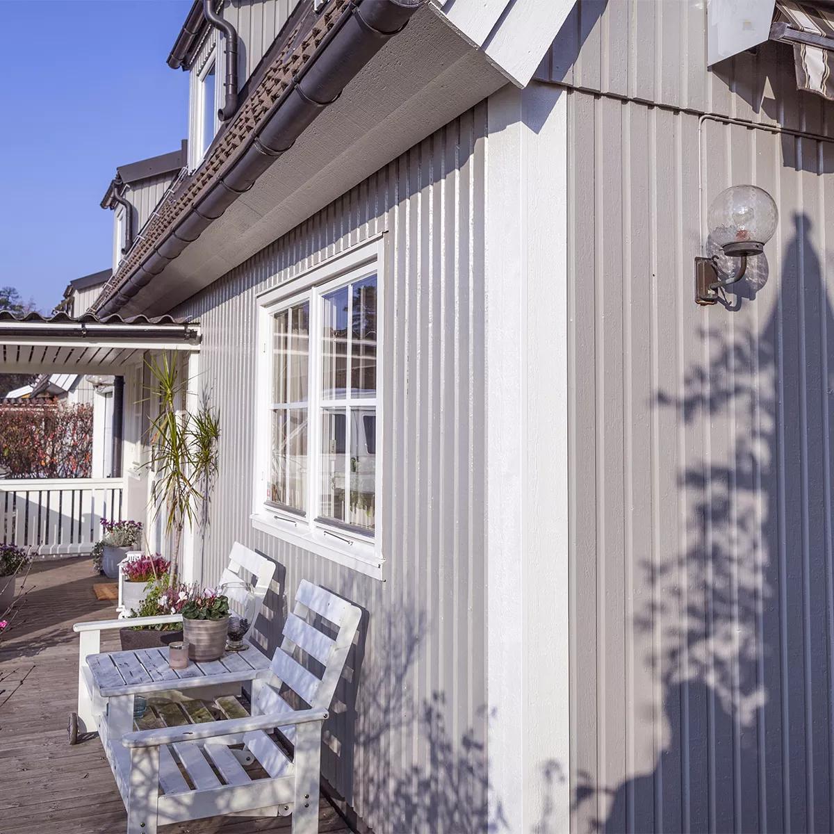 Side of a light grey house with vertical siding, a white-framed window, and a wooden deck with two white chairs.