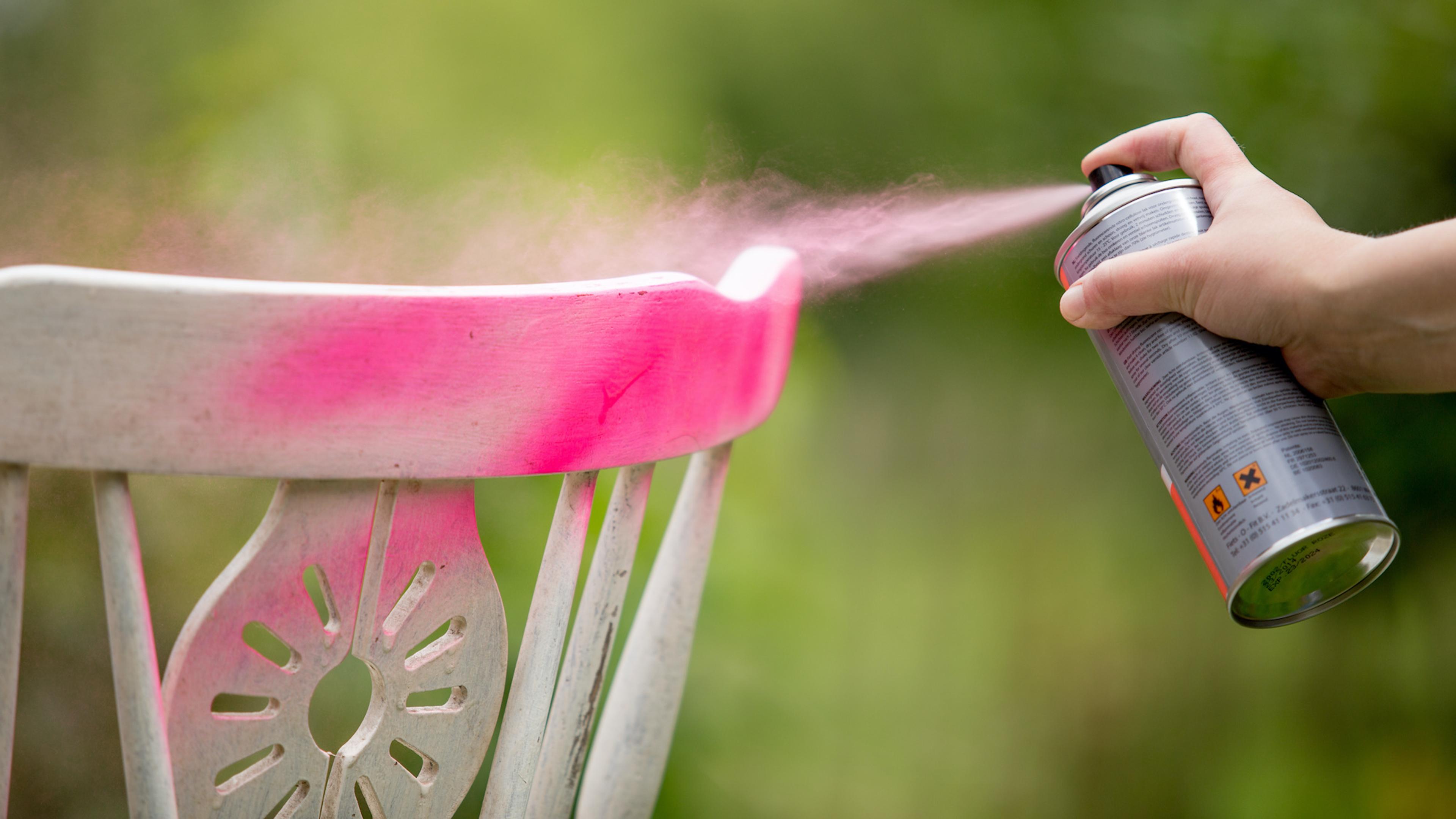 A hand spray paints a white wooden chair with pink paint.