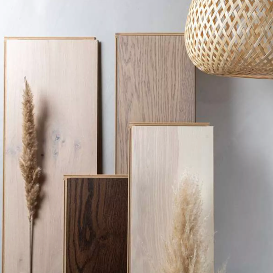 Stacked wood flooring samples in light, medium, and dark tones, with dried pampas grass and a woven light shade.