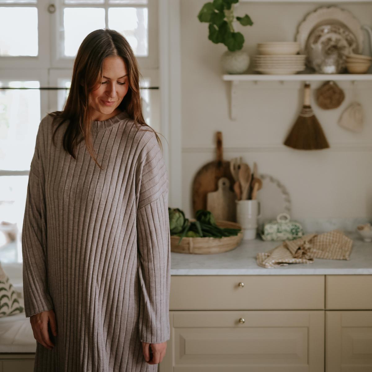 A smiling woman in a long ribbed dress stands in a cozy kitchen.