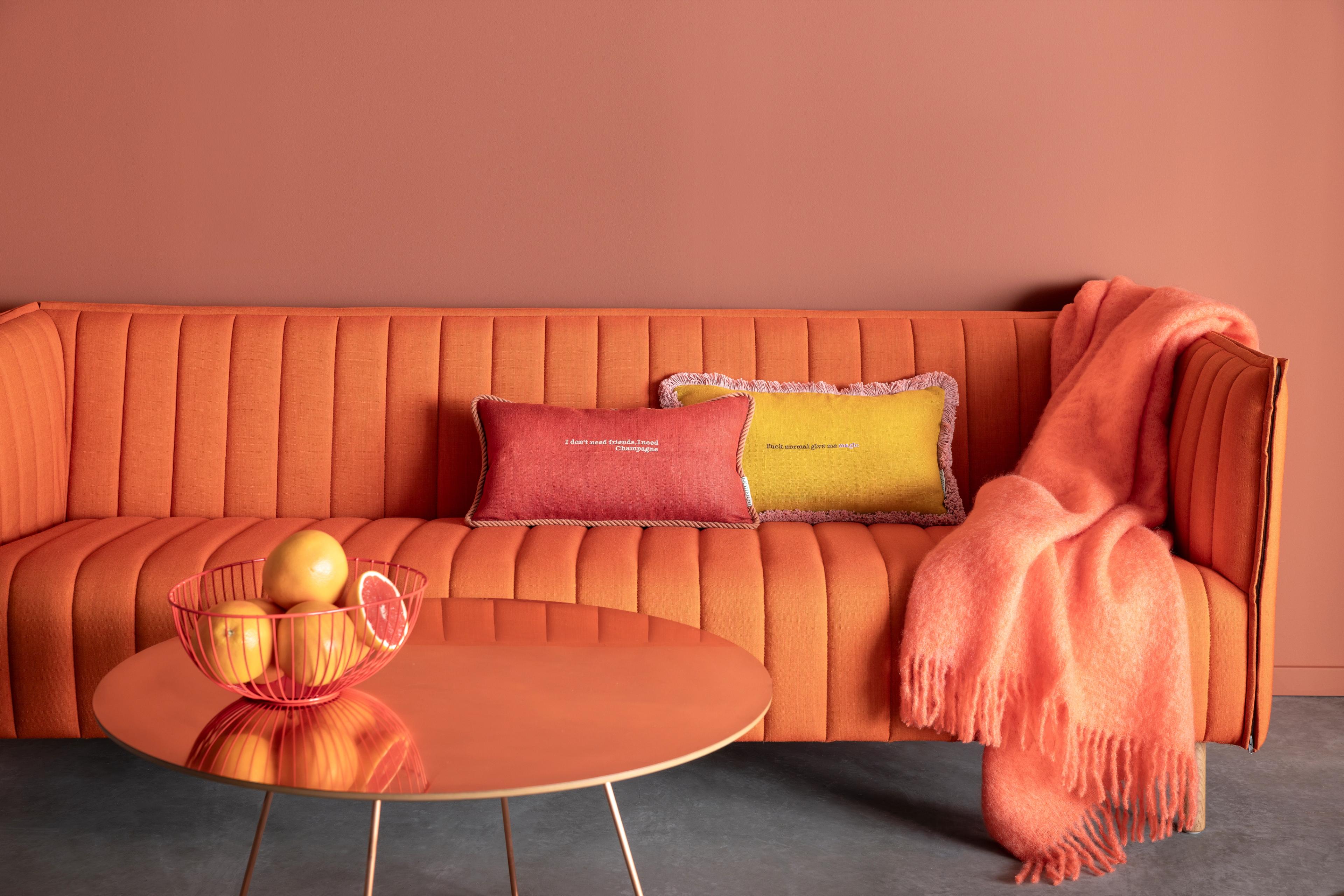 A vibrant living room with an orange sofa, coral-pink walls, a matching coffee table holding a bowl of grapefruits, and a fluffy throw blanket.