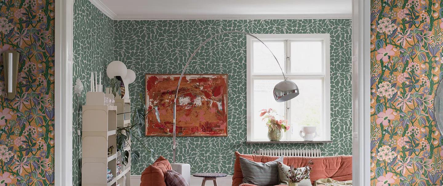 A living room with two distinct wallpapers: a green abstract pattern on the main wall and a colorful floral pattern on the side walls, featuring an orange sofa.