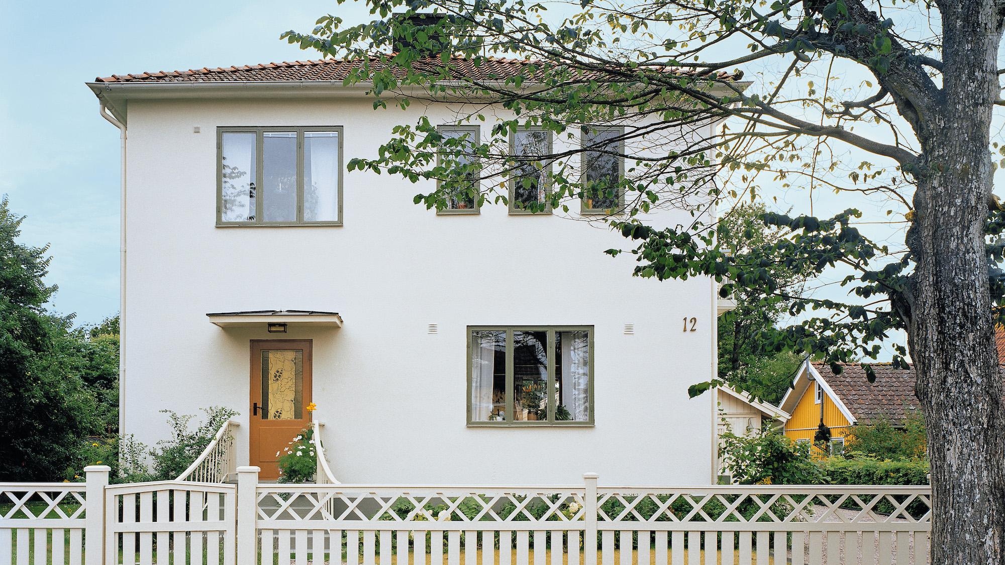 A white two-story house with green windows, a wooden front door, and a white picket fence.