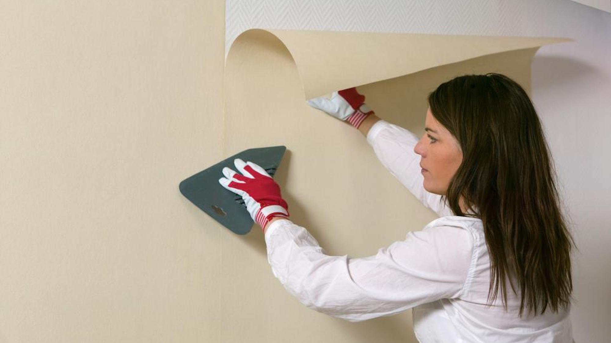 A woman wearing gloves applies beige wallpaper to a wall with a smoothing tool.