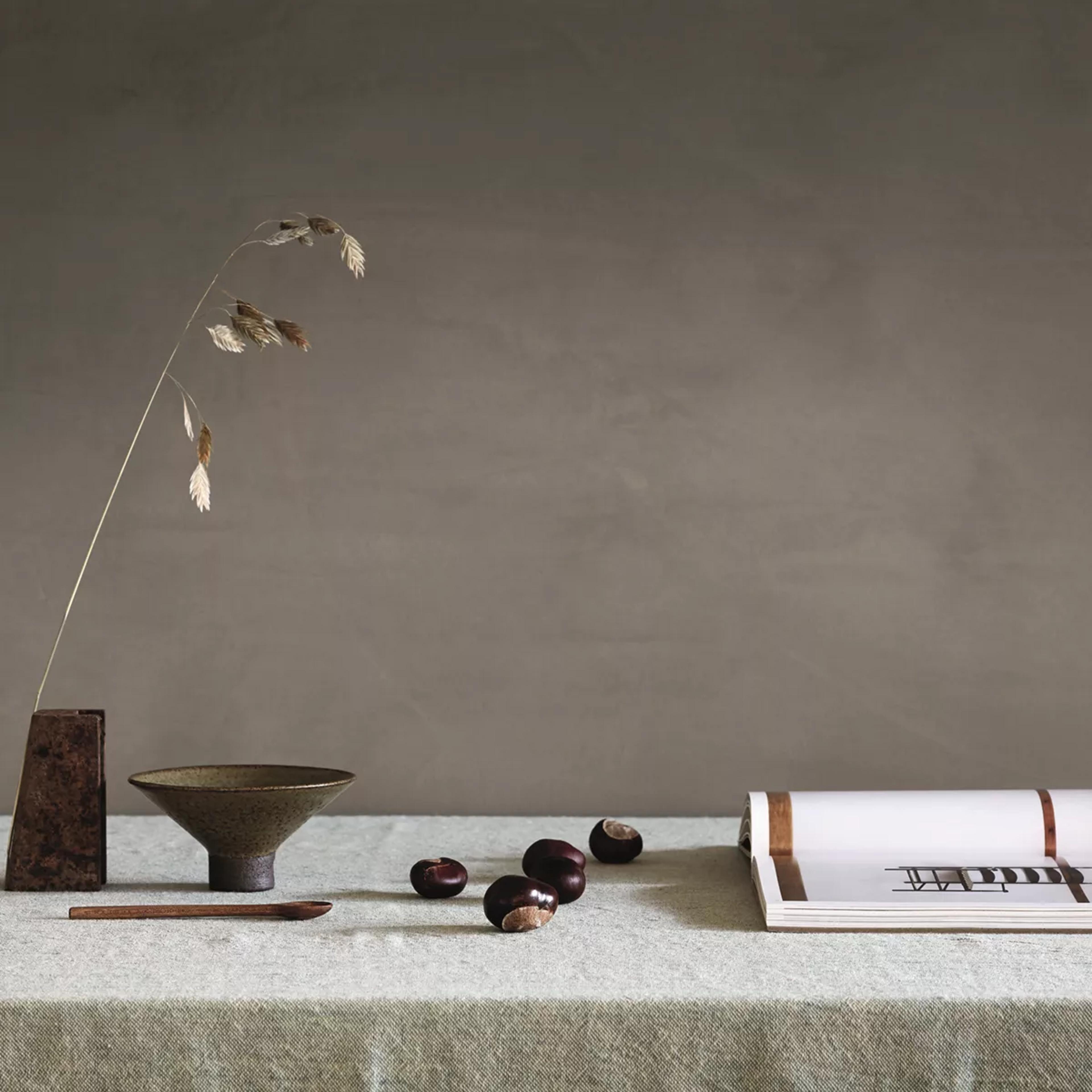 Autumnal still life: dried grass in a vase, ceramic bowl, chestnuts, and an open book on a green linen tablecloth.