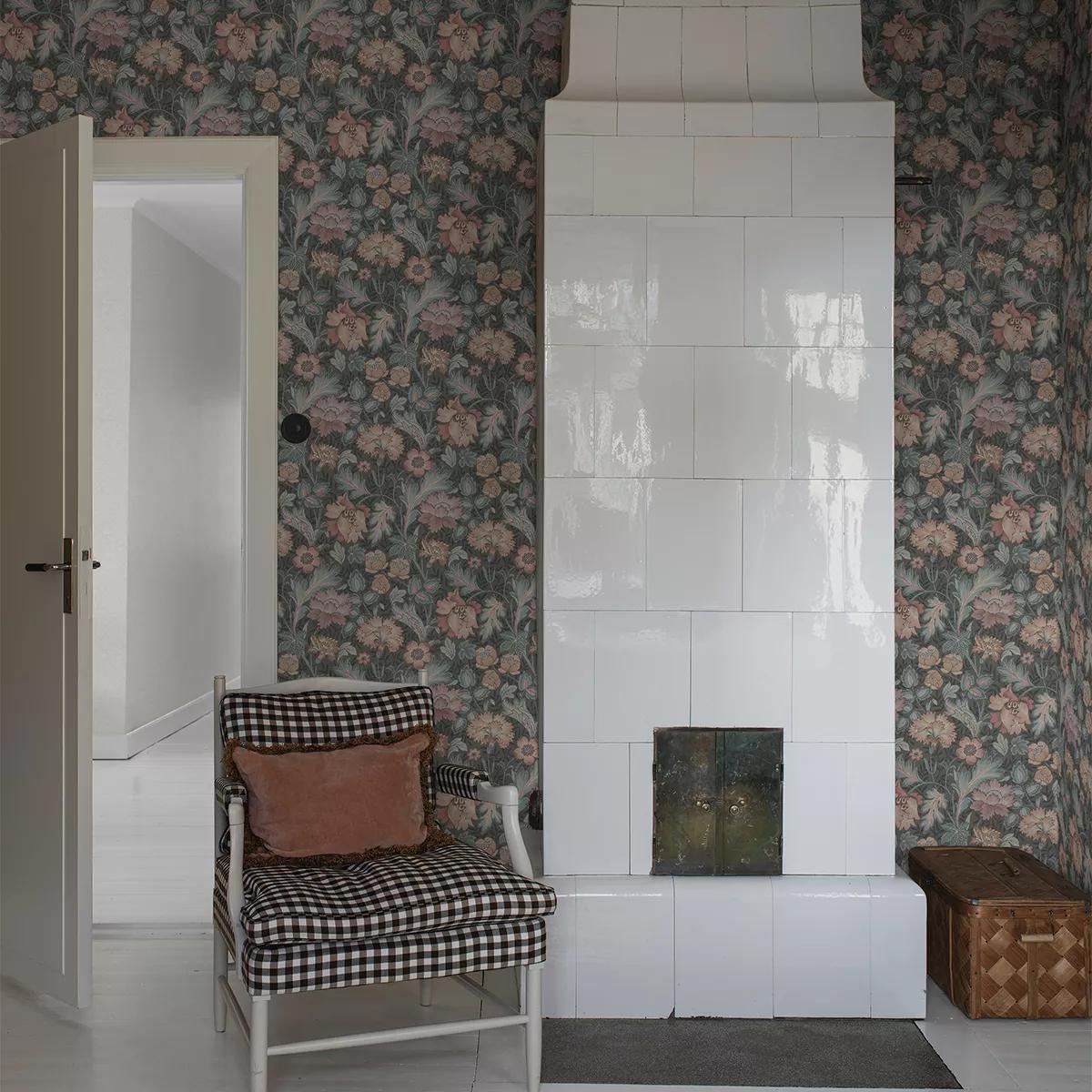 A room with dark floral wallpaper, a tall white tiled fireplace, a black and white gingham armchair with a pink pillow, and an open white door.