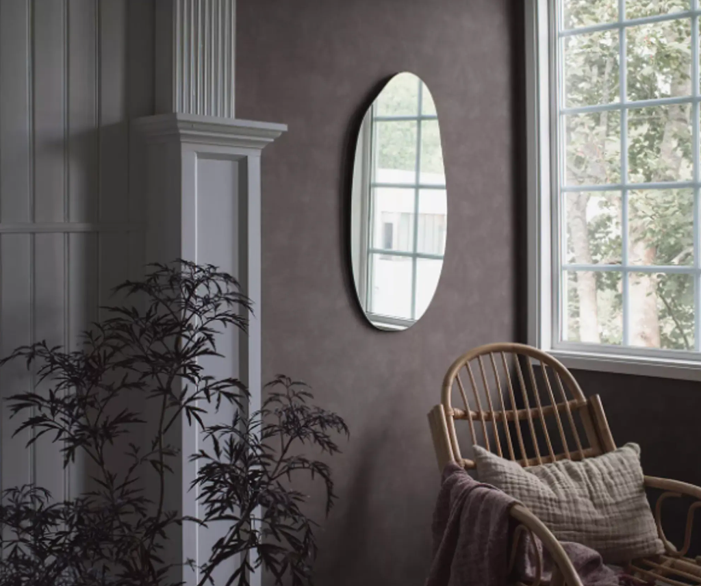 A cozy room with dark plum walls, a white paneled accent, an irregular oval mirror, a rattan chair by a large window, and a dark plant.