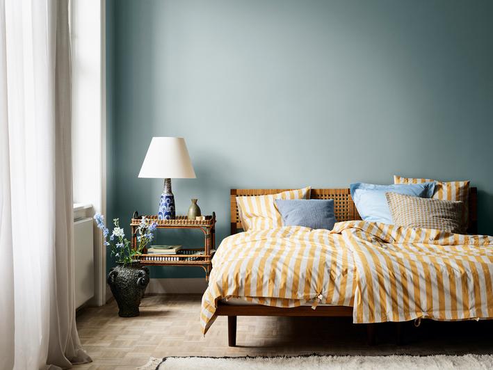 A stylish bedroom with light teal walls, yellow and white striped bedding on a wooden bed, a rattan nightstand, and blue flowers.