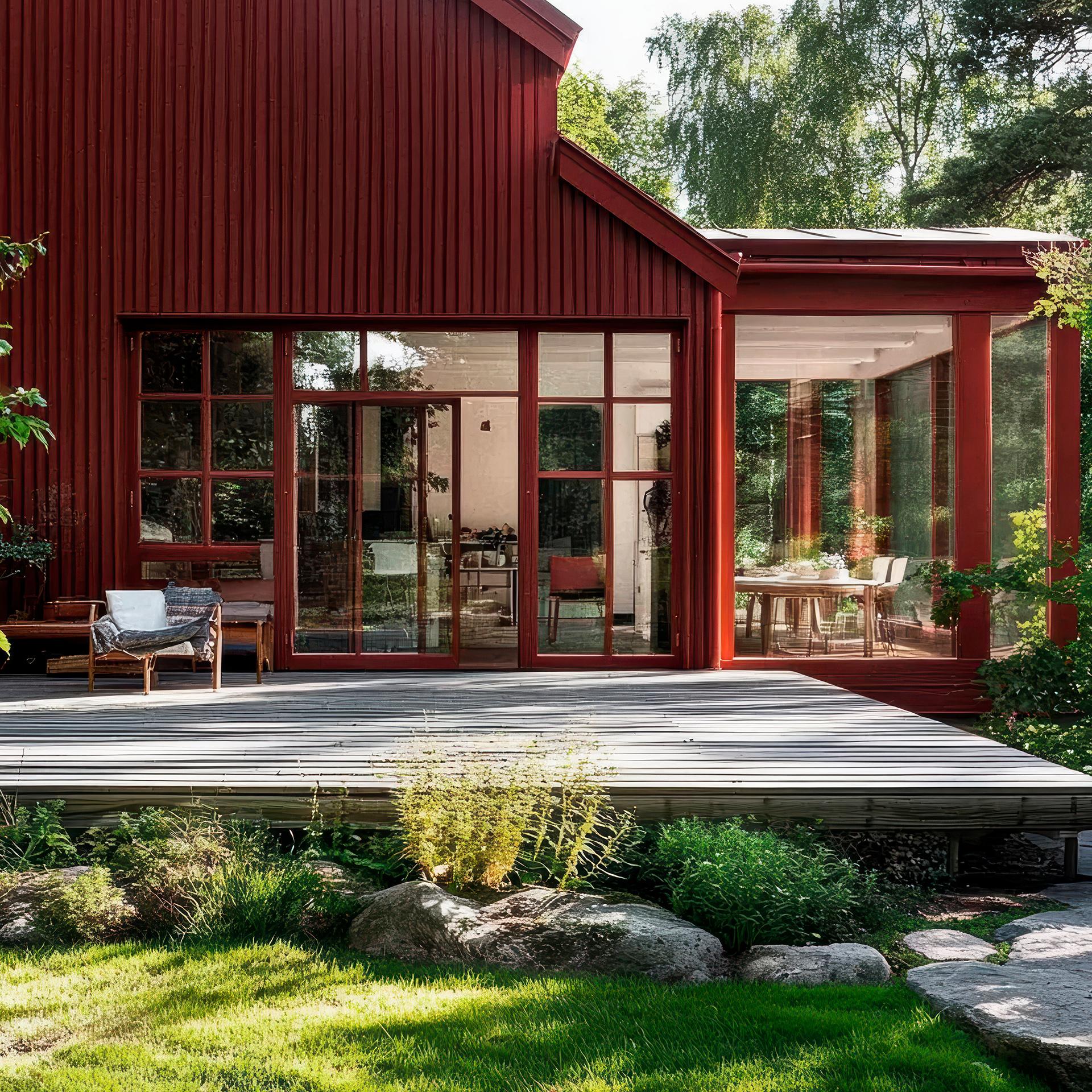 A modern red house with large windows, a wooden deck, and a glass sunroom, set in a lush green garden.