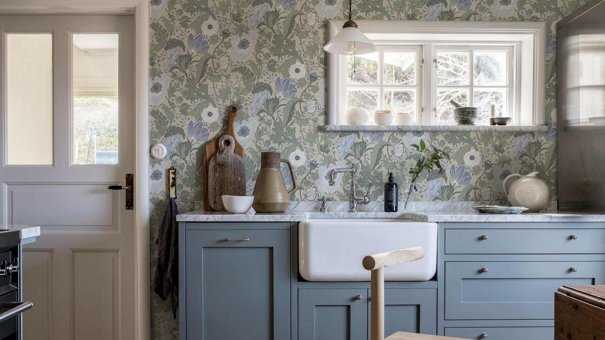 A kitchen with green and blue floral wallpaper, light blue lower cabinets, a white farmhouse sink, and a window.